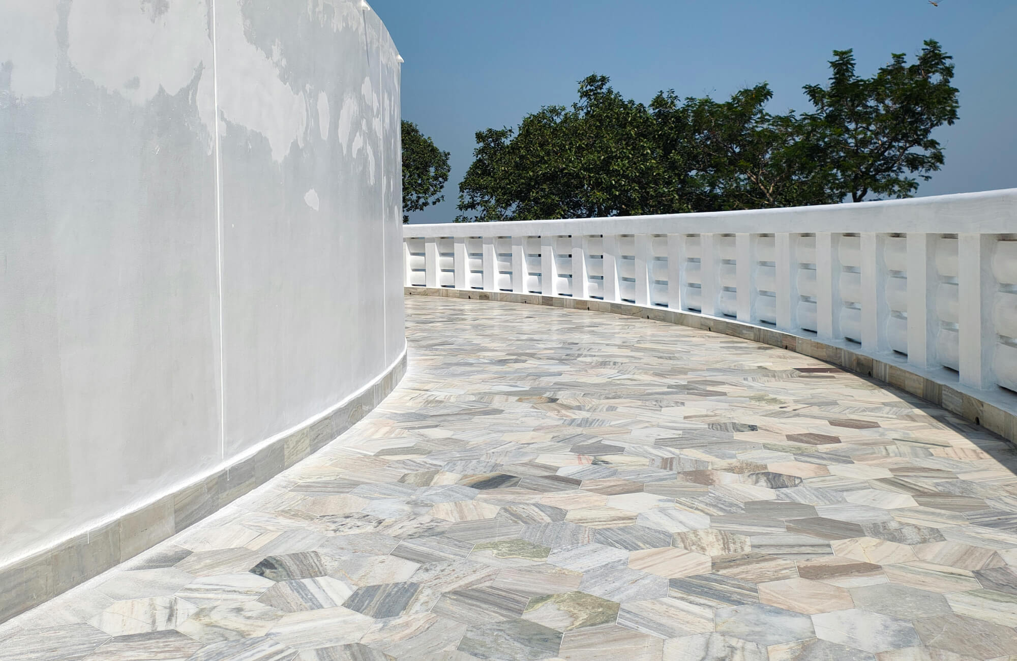 null Curved outdoor terrace with mixed-tone marble mosaic flooring, white railings, and trees under a clear blue sky.