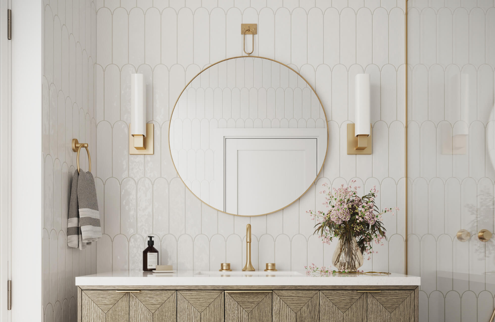 Elegant bathroom with a round brass-framed mirror, gold fixtures, scalloped white tile walls, and a floral arrangement on the vanity.