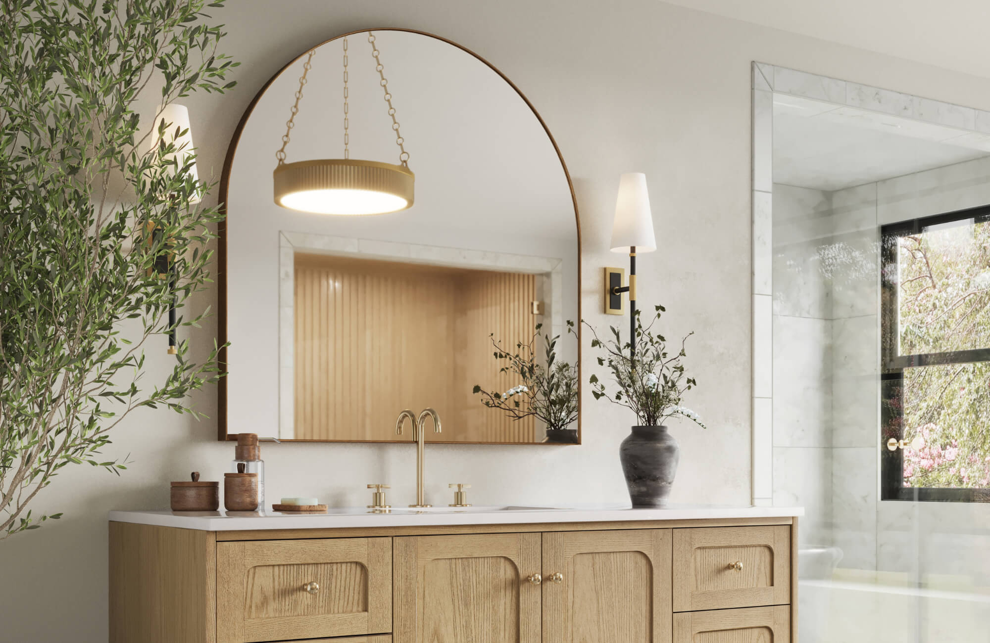 Bathroom with an arched mirror, gold faucet, wood vanity, wall sconces, and greenery beside a large window and walk-in shower.