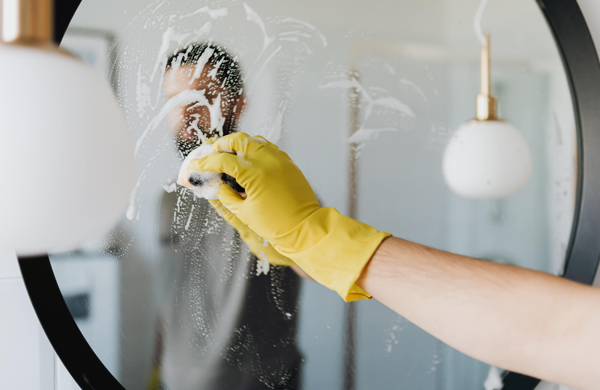 Person wearing yellow gloves cleaning a round mirror with soap and sponge, reflected in the glass under soft pendant lighting.