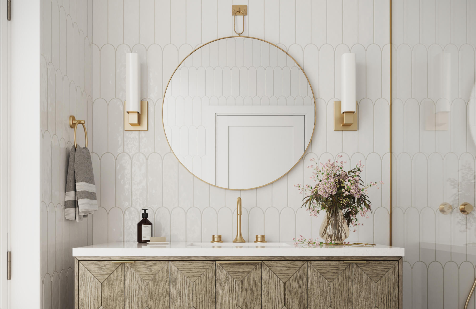 Elegant bathroom with a round gold-framed mirror, wall sconces, wood vanity, and fresh flowers, styled in a soft, modern neutral palette.