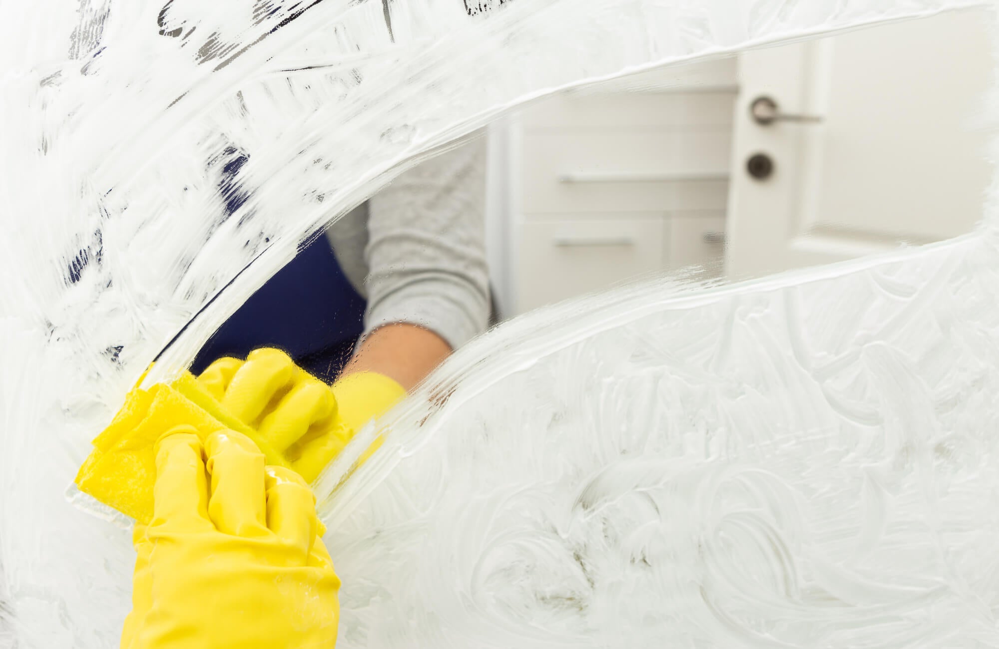 A person wearing yellow gloves is cleaning a mirror with a sponge, revealing a clear reflection amid white cleaning solution in a bright room.