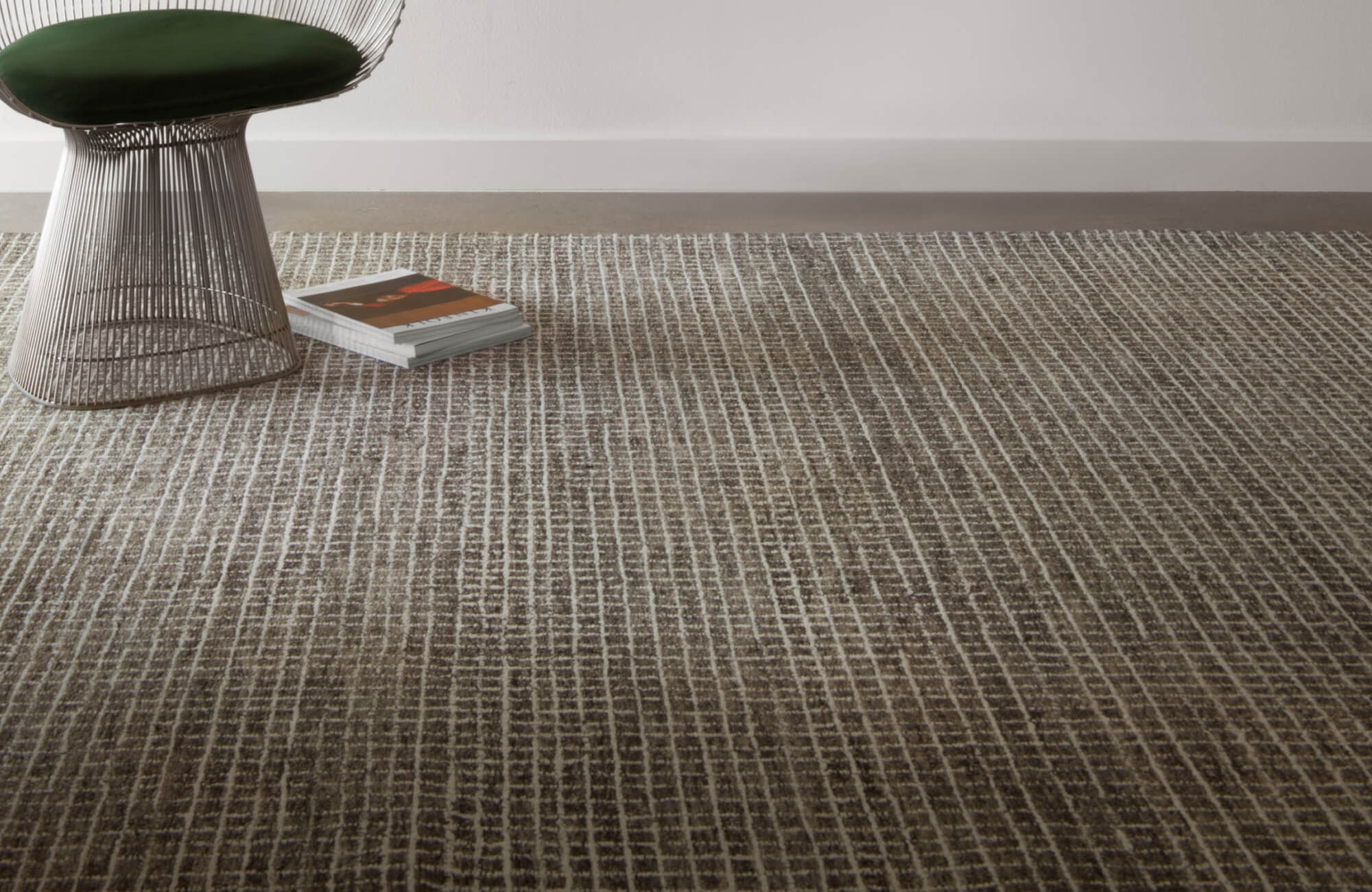 Modern wool area rug with a linear grid pattern in earthy tones, paired with a sculptural chair and stacked books.