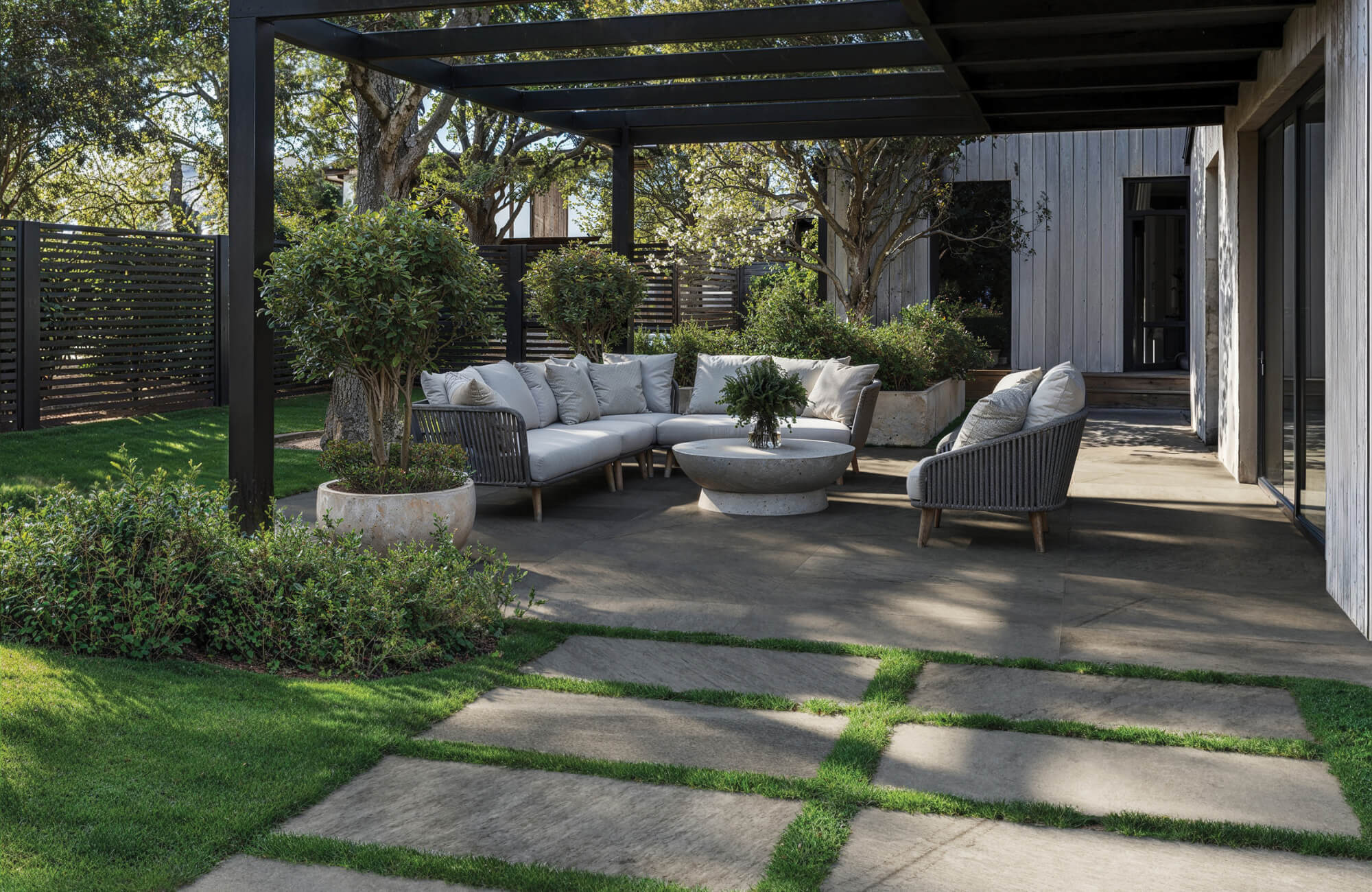 Modern outdoor patio with large gray porcelain pavers, cushioned seating under a black pergola, and lush greenery surrounding the space.