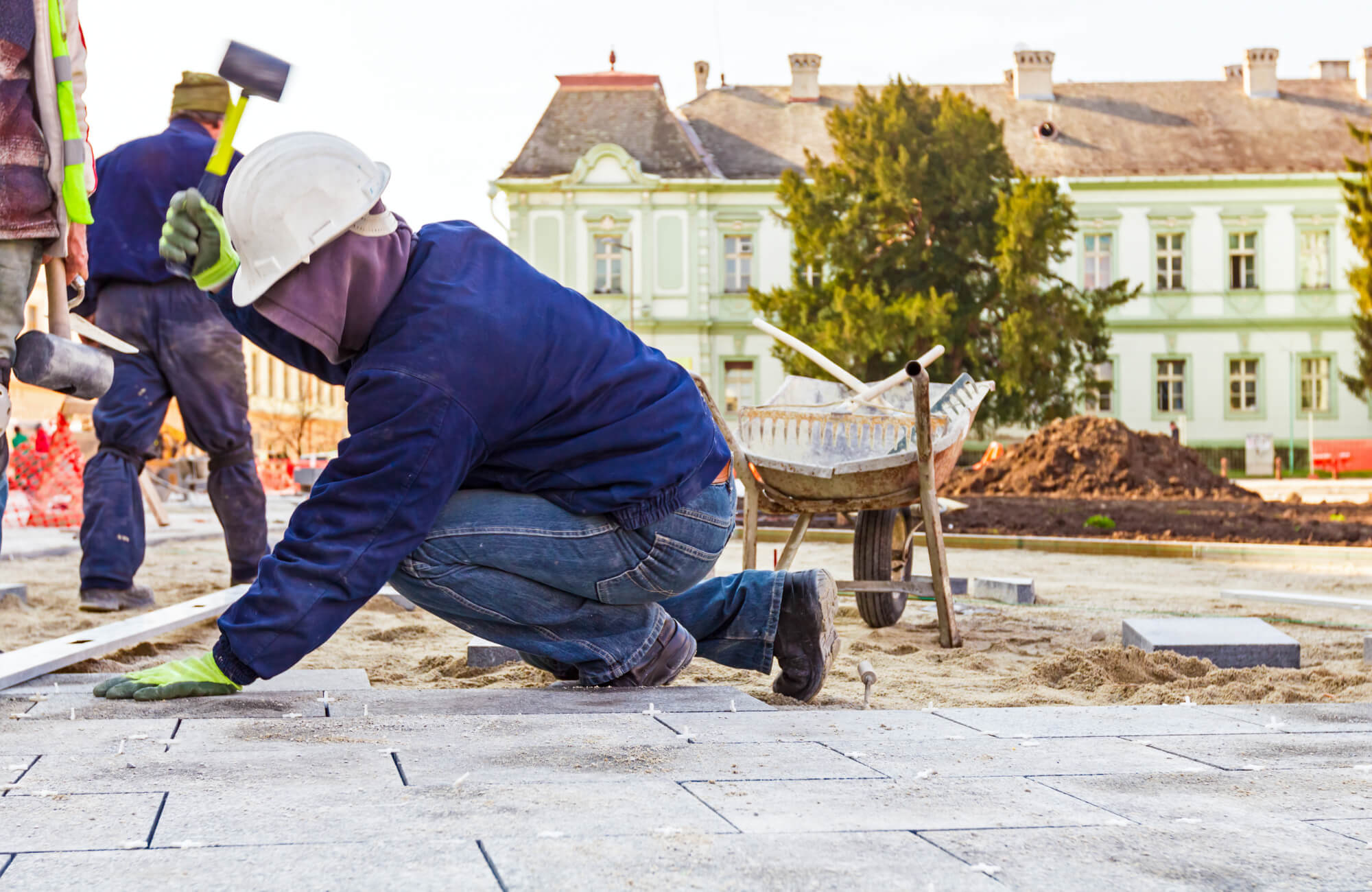 Construction workers installing outdoor pavers on a prepared sand base, with tools and a wheelbarrow on-site near a historic building.