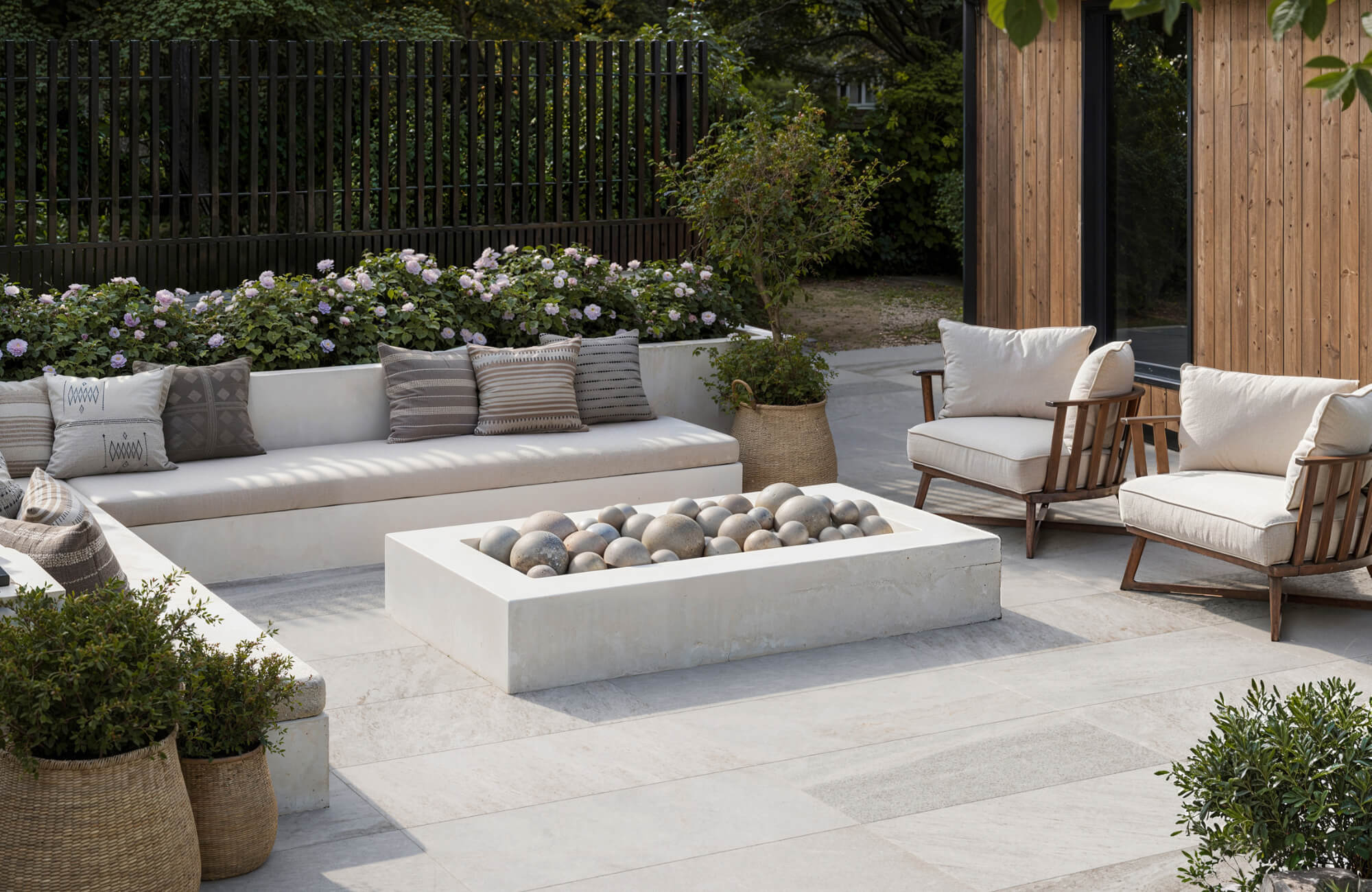 Stylish outdoor lounge with light porcelain pavers, built-in seating, cushioned chairs, and a modern fire feature surrounded by greenery.