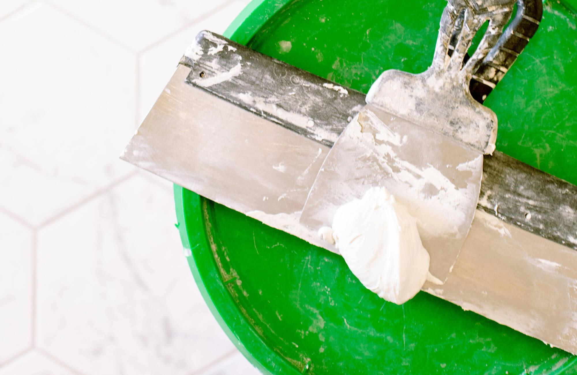 Close-up of a trowel with grout on a bright green bucket, ready for tile application on a hexagonal tile surface.