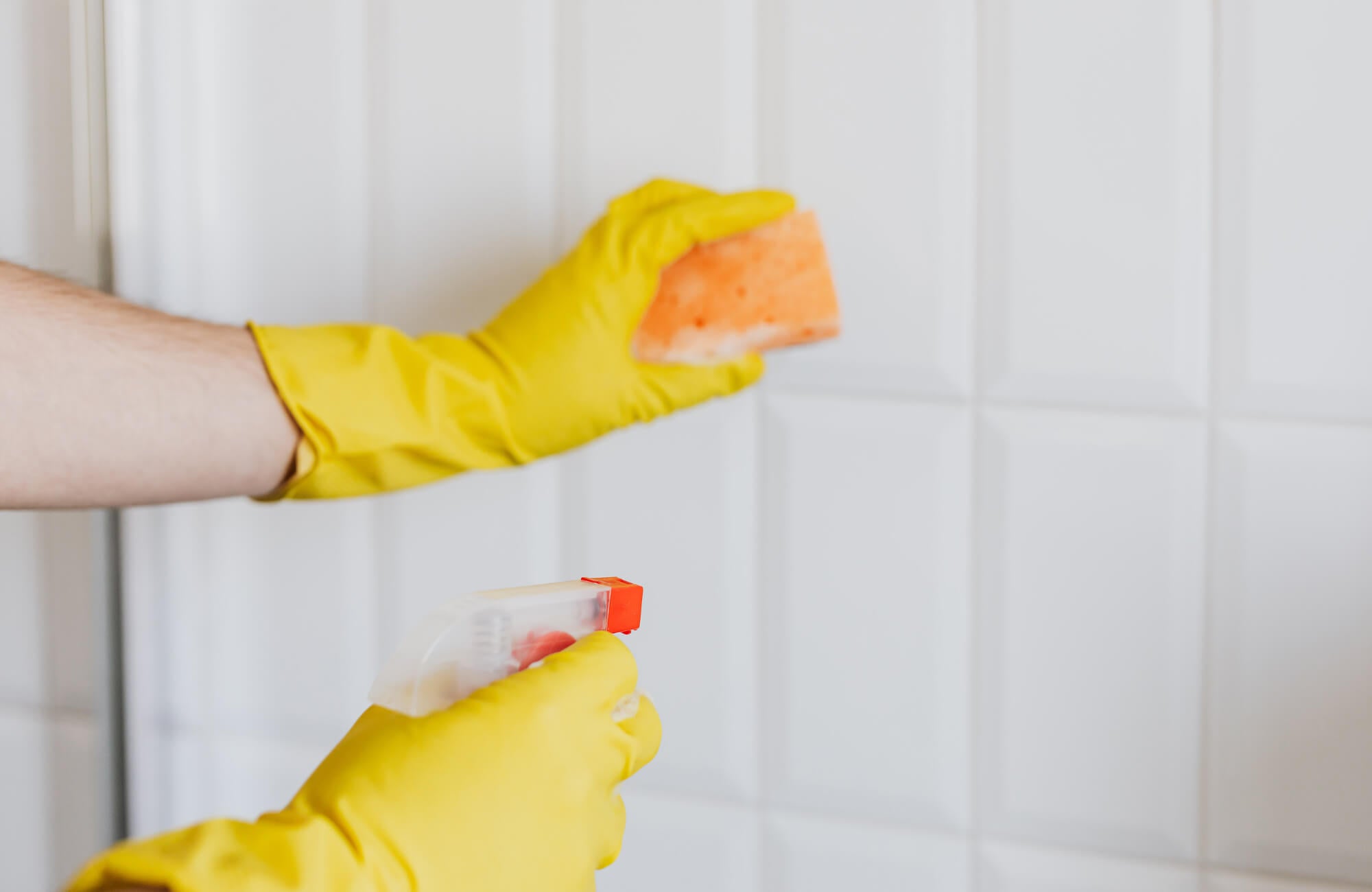 null Person wearing yellow rubber gloves cleaning white tiled backsplash with an orange sponge and spray bottle.