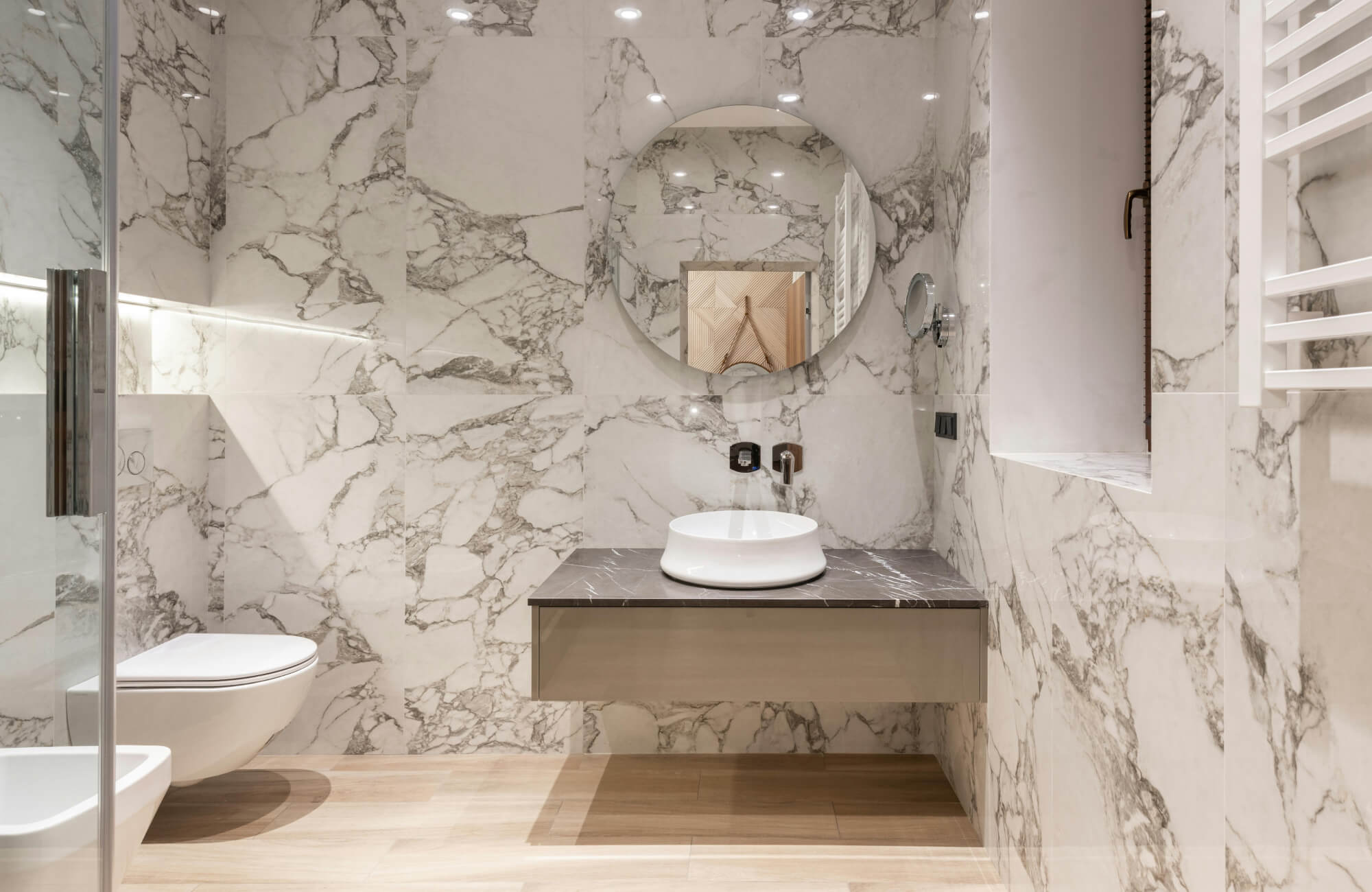 null Floating vanity with marble countertop, round mirror, and vessel sink against full marble walls in a modern bathroom.