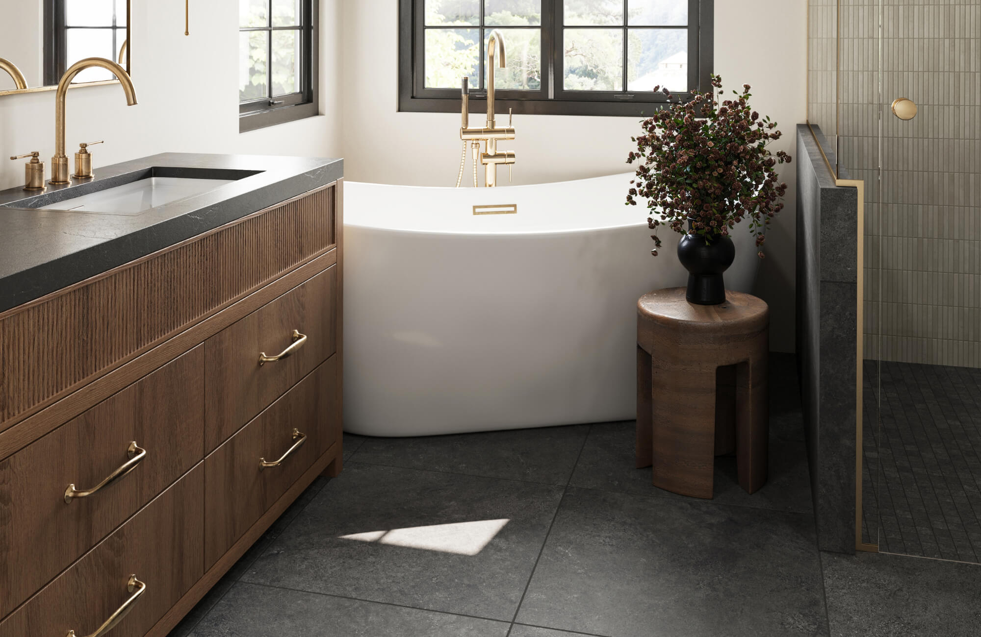 Modern bathroom with matte black floor tiles, freestanding white tub, wood vanity with brass hardware, and natural light from black-framed windows.