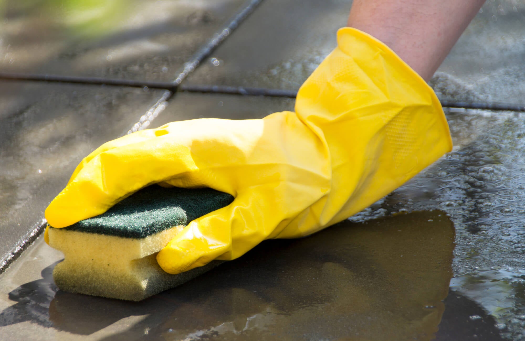 Close-up of a hand in a yellow glove scrubbing wet black tiles with a sponge, highlighting tile cleaning and maintenance in progress.