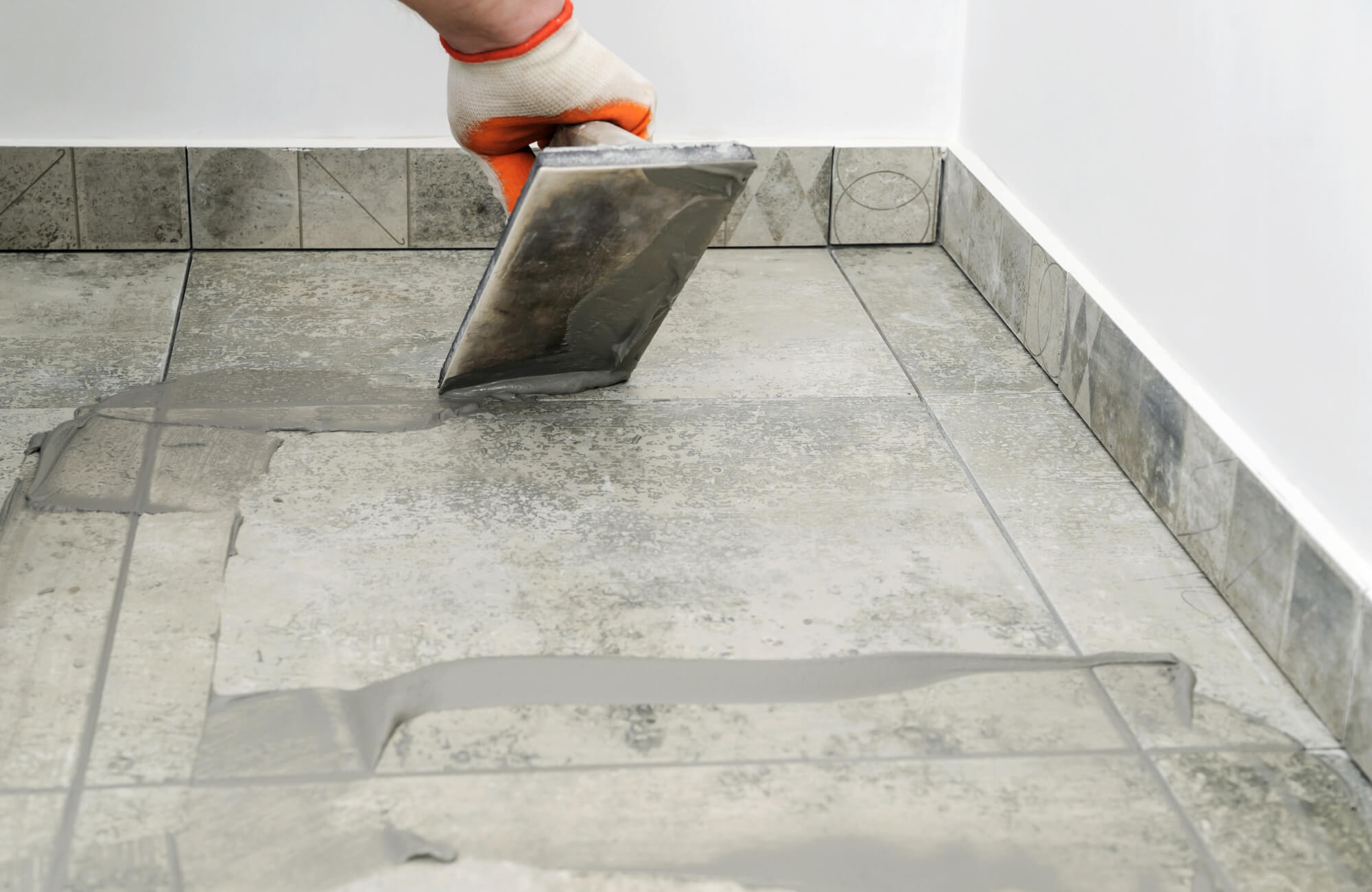 Tile installer applying gray mortar across floor tiles with a trowel during a professional installation process.