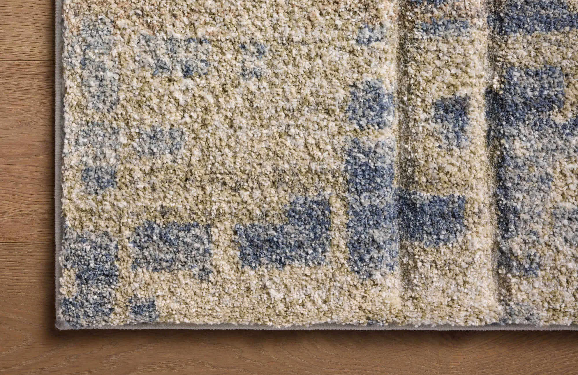 Close-up of a thick, textured rug corner on a warm wood floor, showing an abstract beige, taupe, and muted blue pattern with a bound edge.