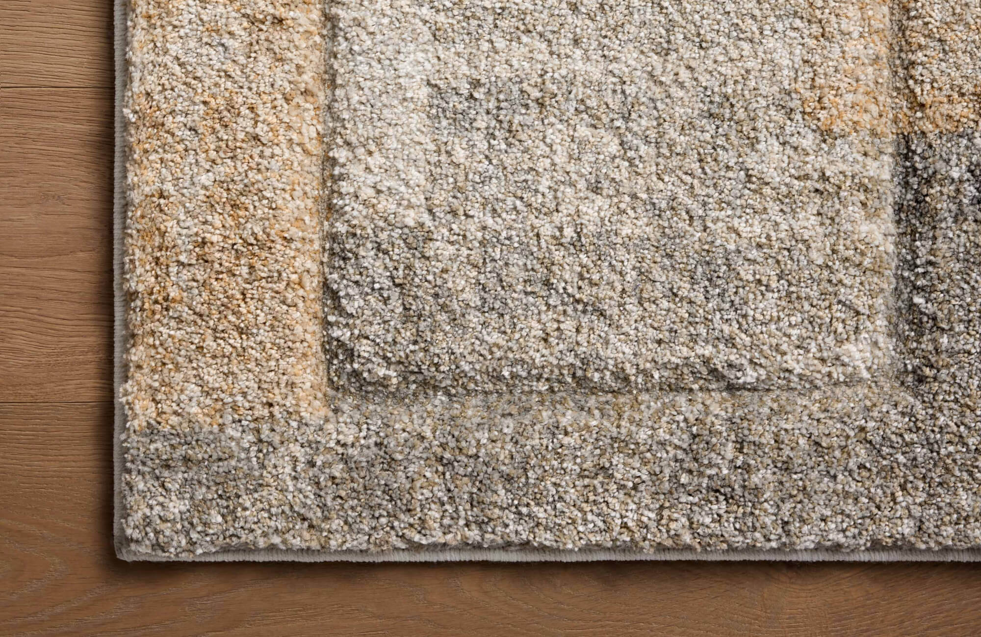 null Close-up of a plush rug corner on a wood floor, featuring a raised geometric border pattern in soft gray, cream, beige, and warm tan tones.