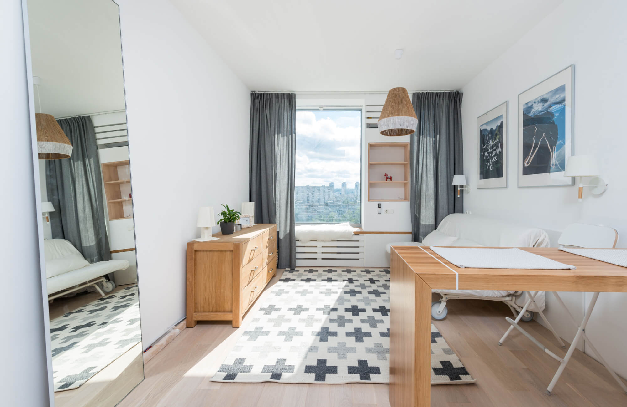 null Bright compact bedroom with a gray-and-white patterned rug under a wooden desk and bed area, framed by a large window, gray curtains, wall art, and light wood furniture.