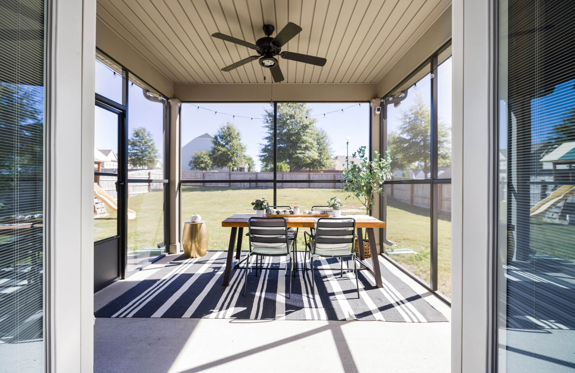 null Covered screened patio with a black-and-white striped rug beneath a wooden dining table, surrounded by glass panels, outdoor chairs, string lights, and a ceiling fan.