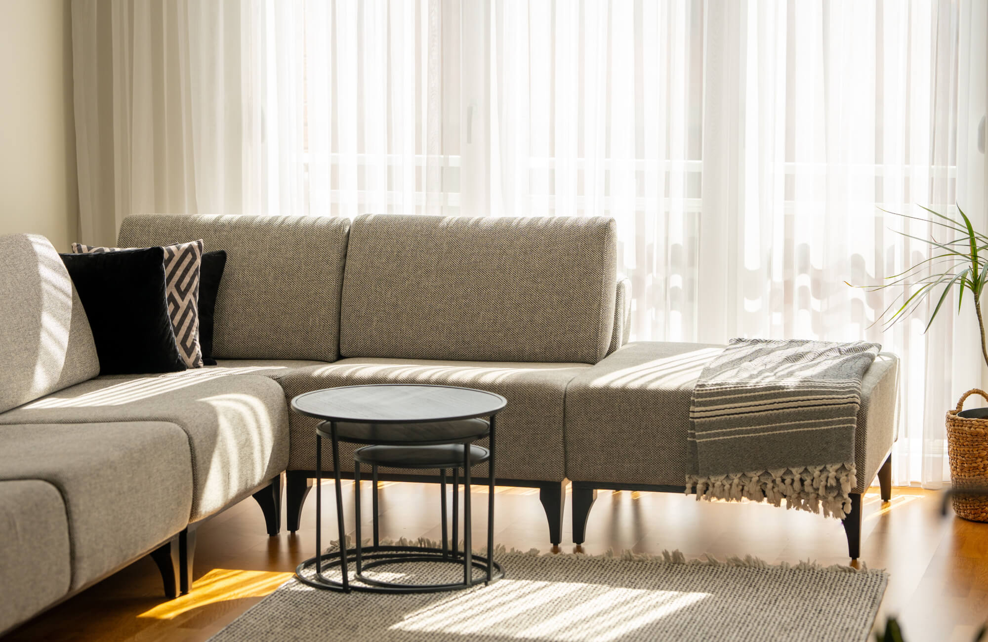 null Sunlit living room with a neutral woven rug under a round nesting coffee table, placed beside a beige sectional sofa, sheer curtains, and a potted plant.