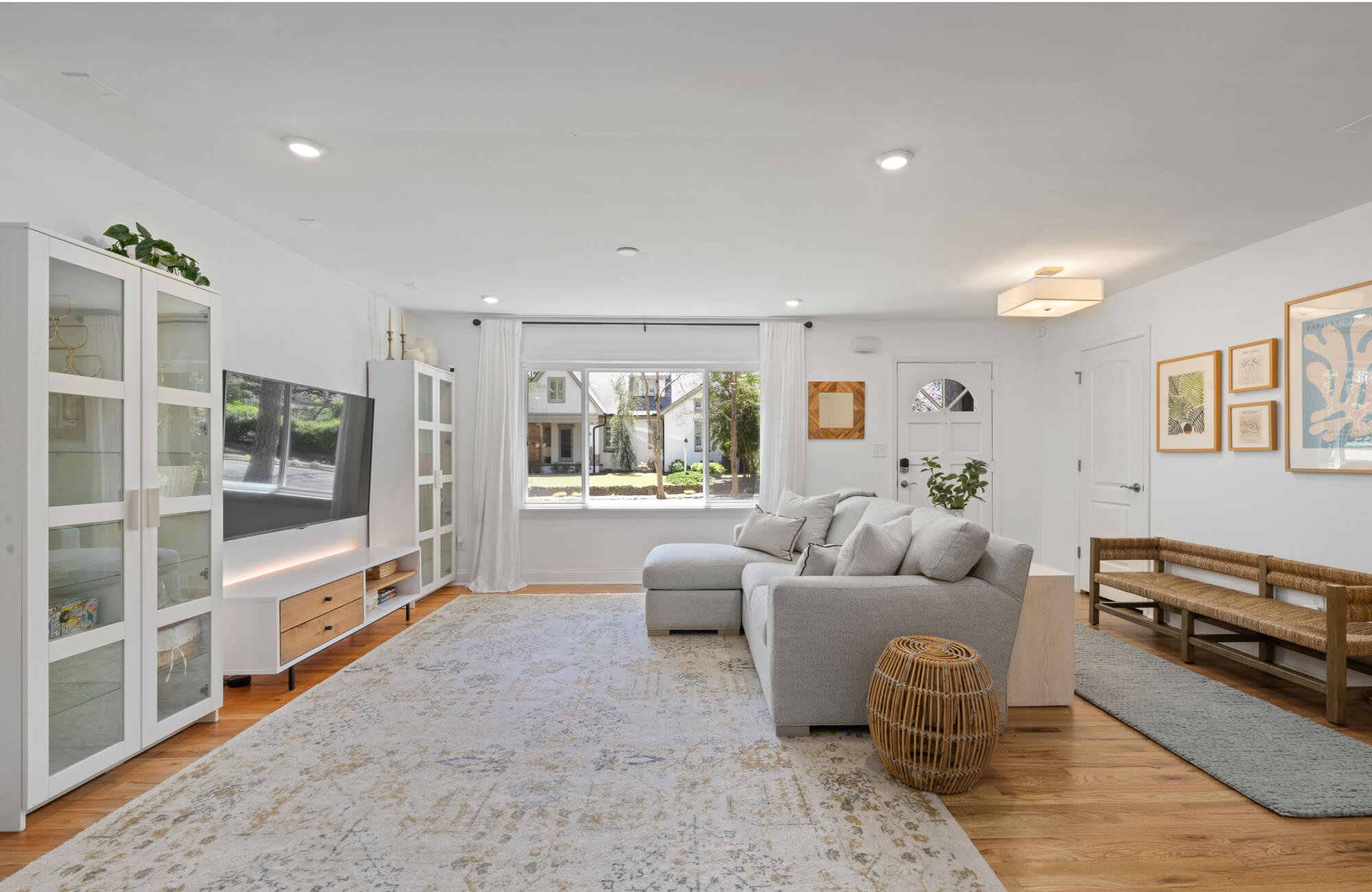 null Open living room with a large light-toned patterned rug under a gray sectional sofa, white media cabinets, wood flooring, framed wall art, and natural light from a front window.