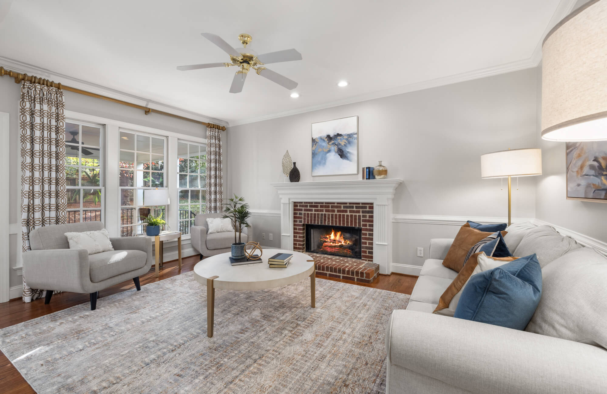 null Traditional living room with a pale distressed rug beneath a round coffee table, positioned between neutral seating, a brick fireplace, tall windows, patterned curtains, and a ceiling fan.