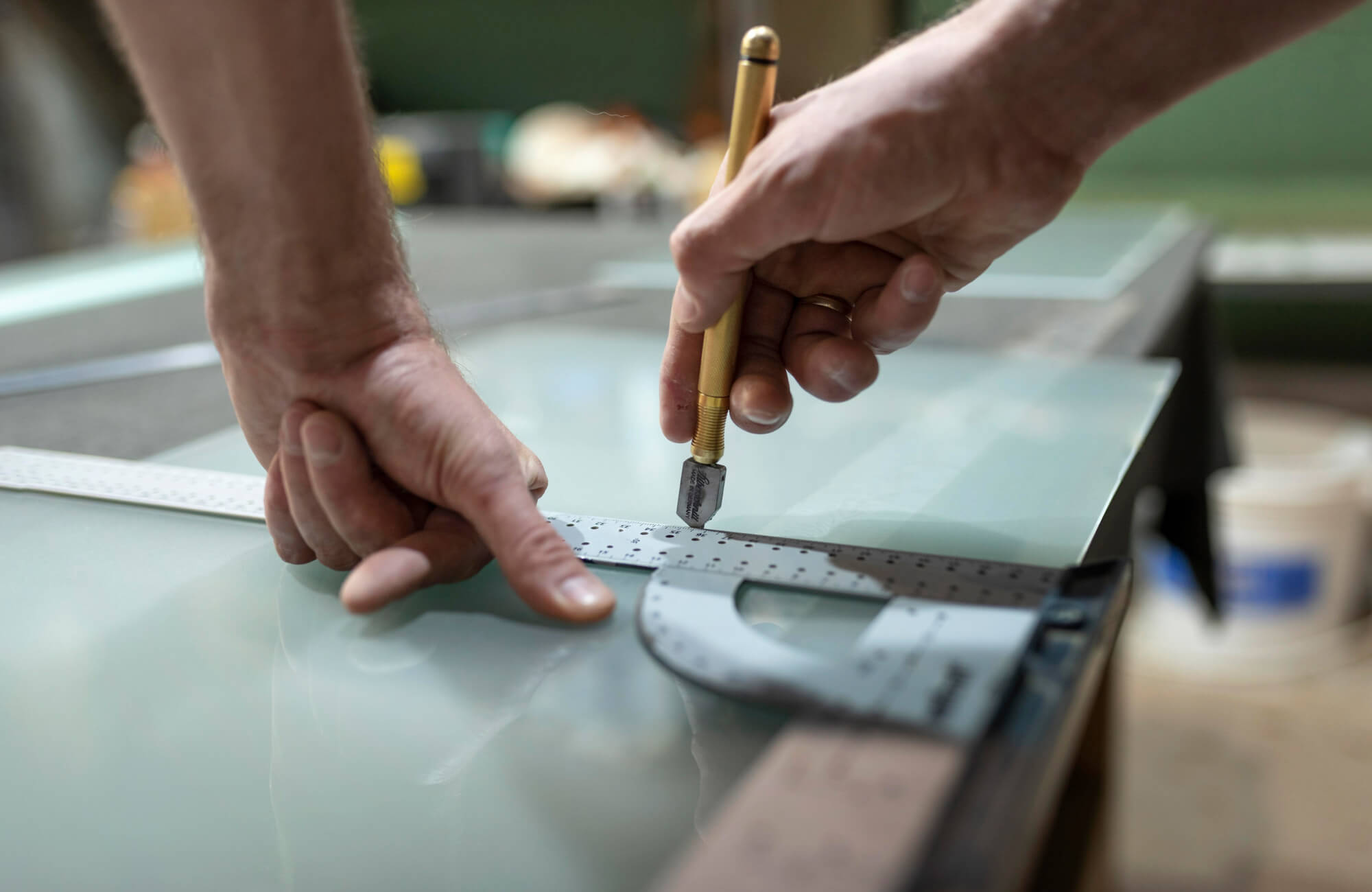 Close-up of a craftsman scoring a glass sheet with a precision cutter and ruler while preparing material for mirror production.