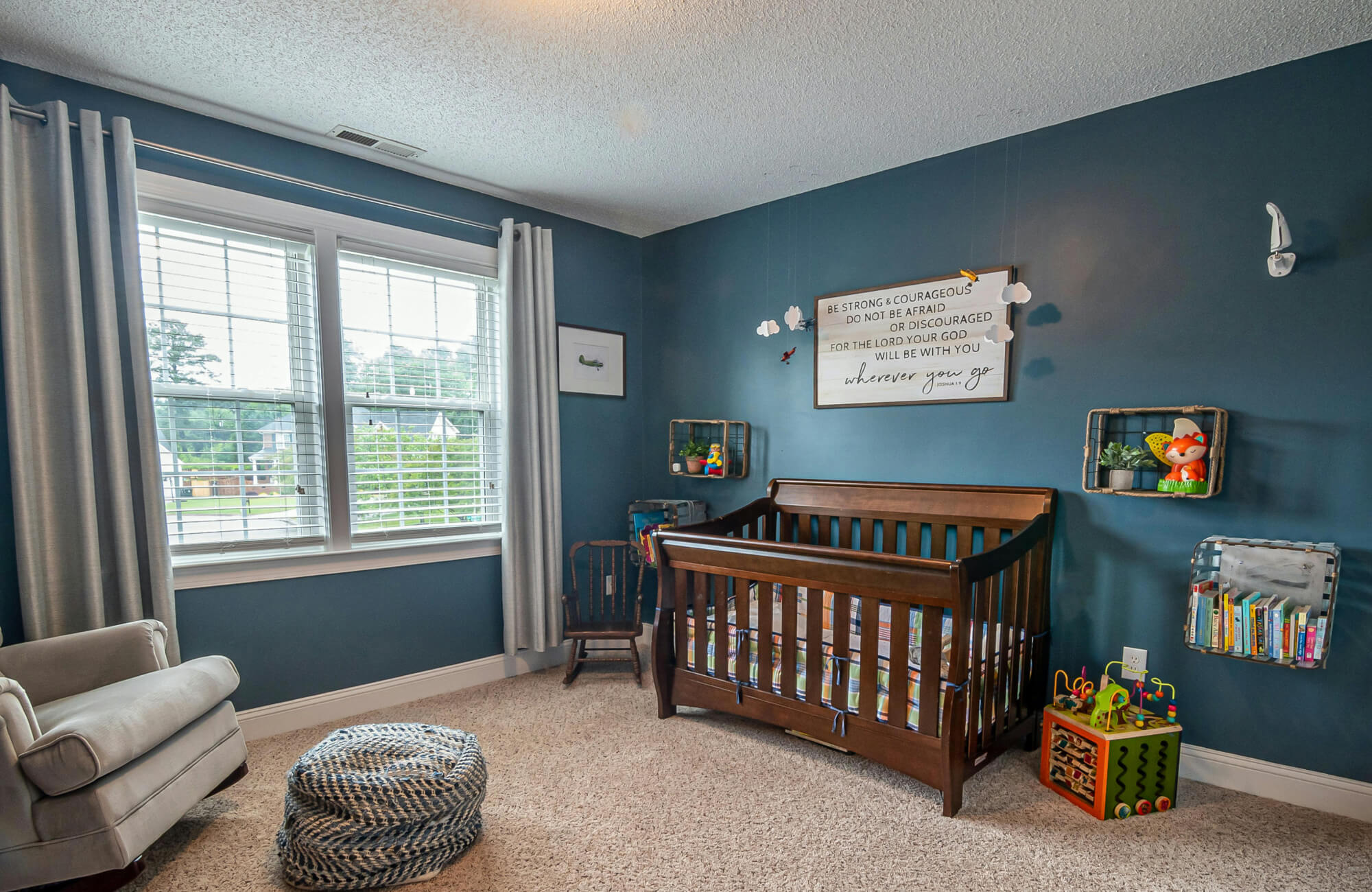 Deep blue nursery with carpeted floor and central rug area that visually anchors the crib and seating arrangement.