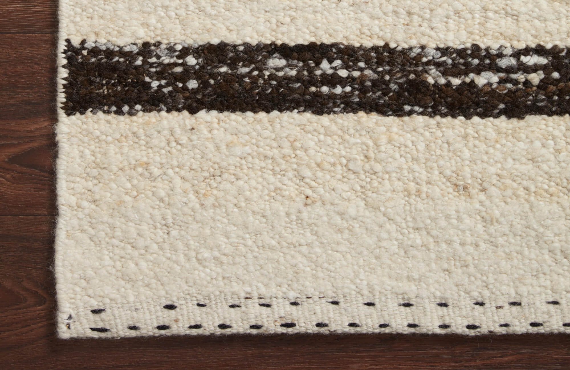 Close-up of a handwoven wool rug with a cream base, dark brown stripe, and subtle stitched border detail.