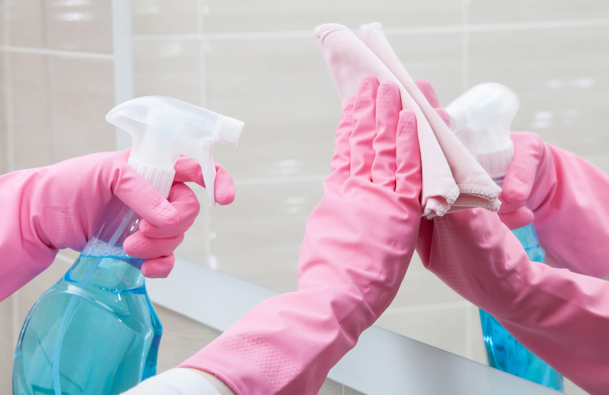 Hands in pink gloves cleaning a mirror with blue glass cleaner and a microfiber cloth to achieve a streak-free reflective surface.