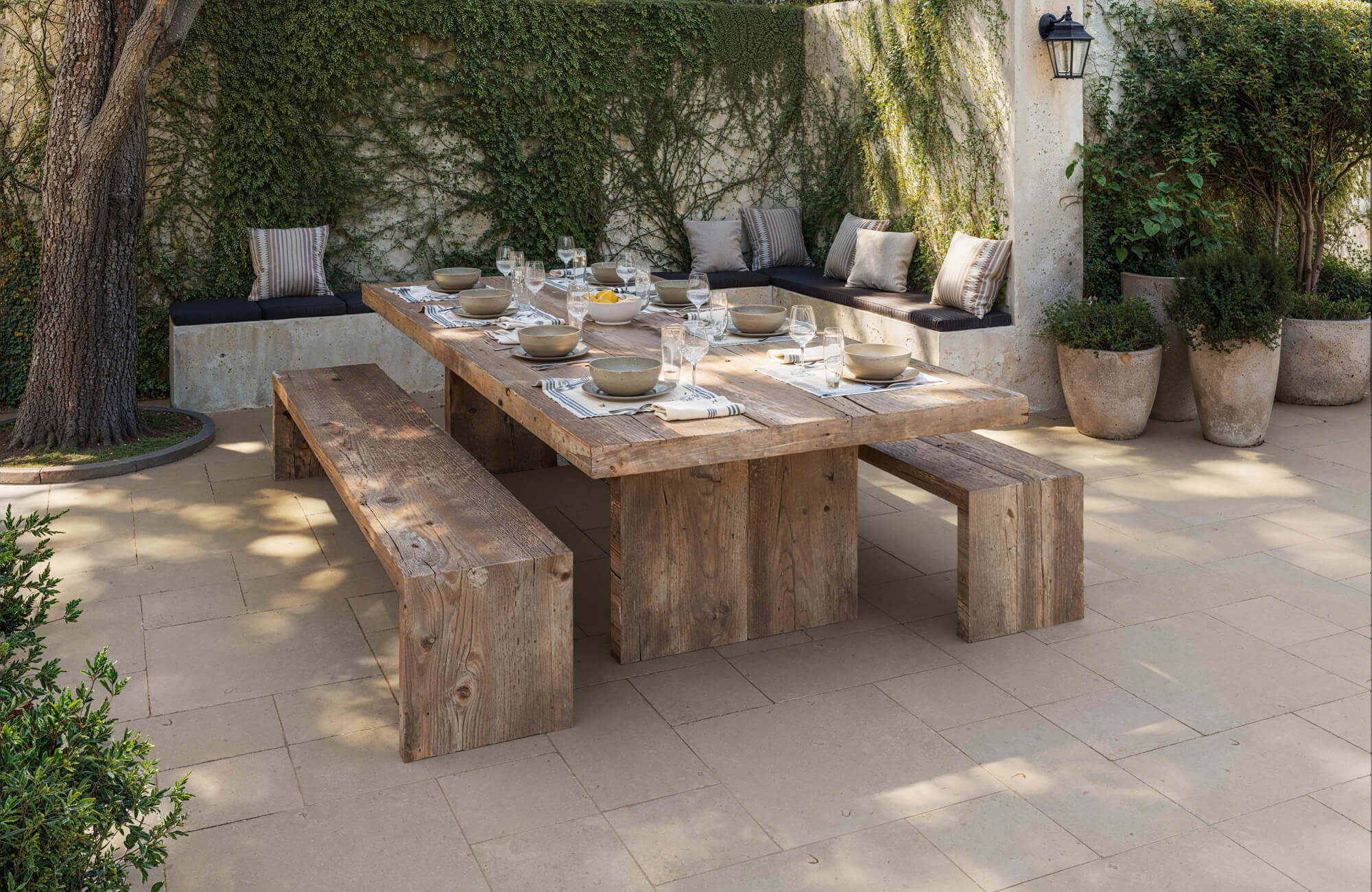 Beige porcelain tiles are laid in a modular pattern beneath a weathered wood table and benches, creating a cozy garden dining setup.