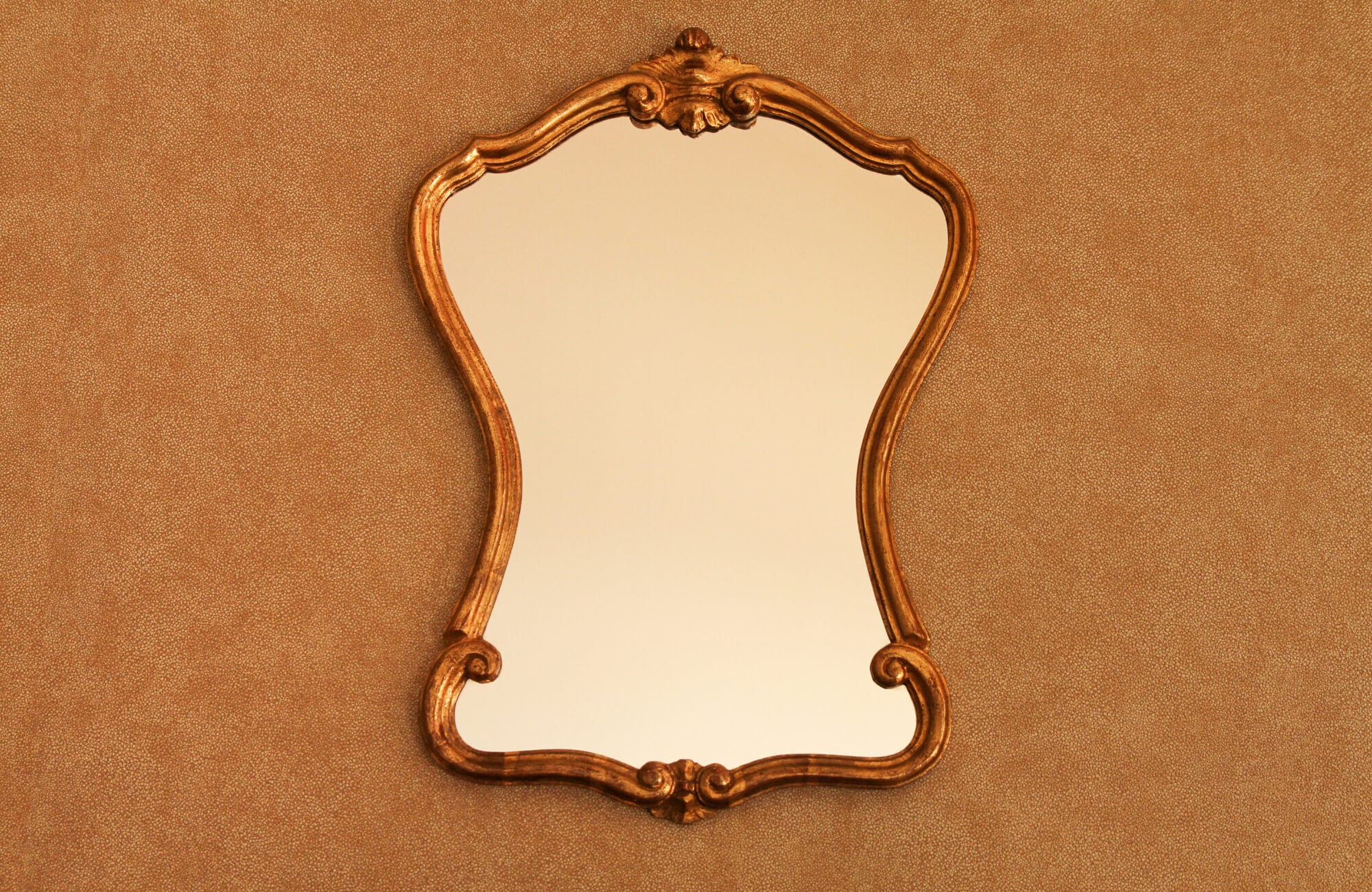 null Ornate vintage wall mirror with a gold carved frame and curving silhouette, displayed against a textured brown background.