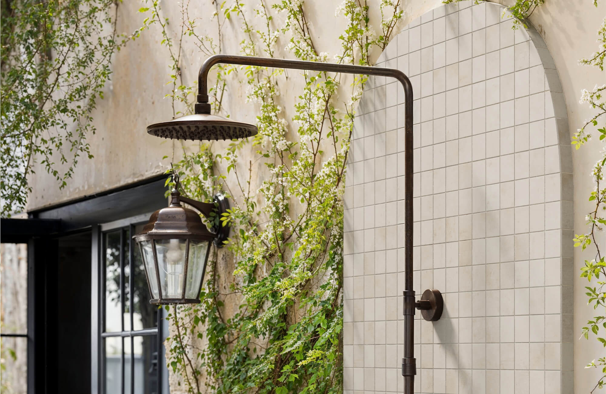 null Charming outdoor shower with vintage bronze fixture and cement look mosaic tiles, framed by climbing greenery and rustic walls.