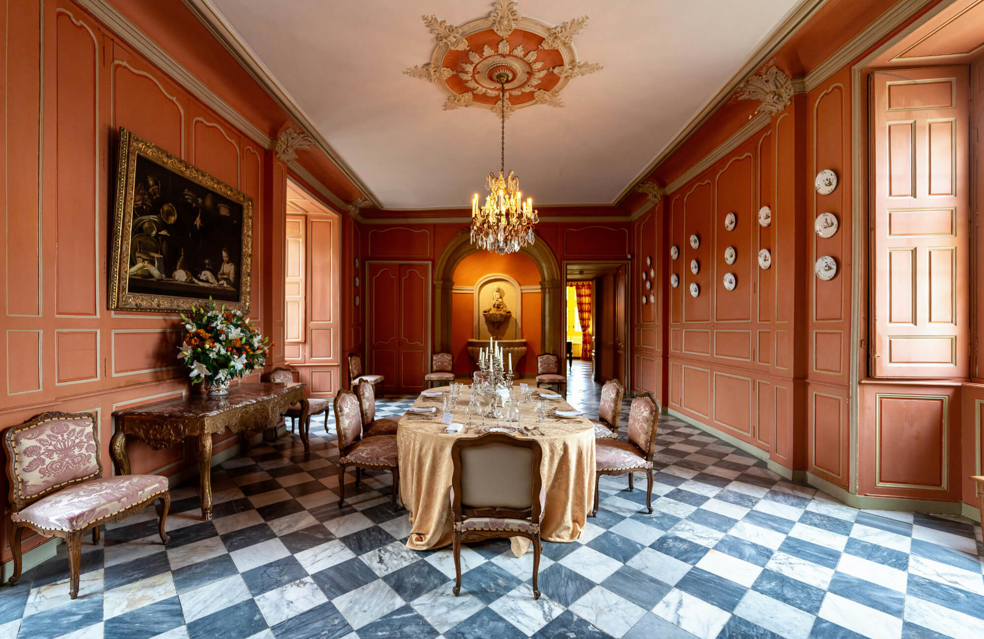 Elegant dining room with a black-and-white marble checkerboard floor, ornate coral-paneled walls, a crystal chandelier, and vintage furnishings.