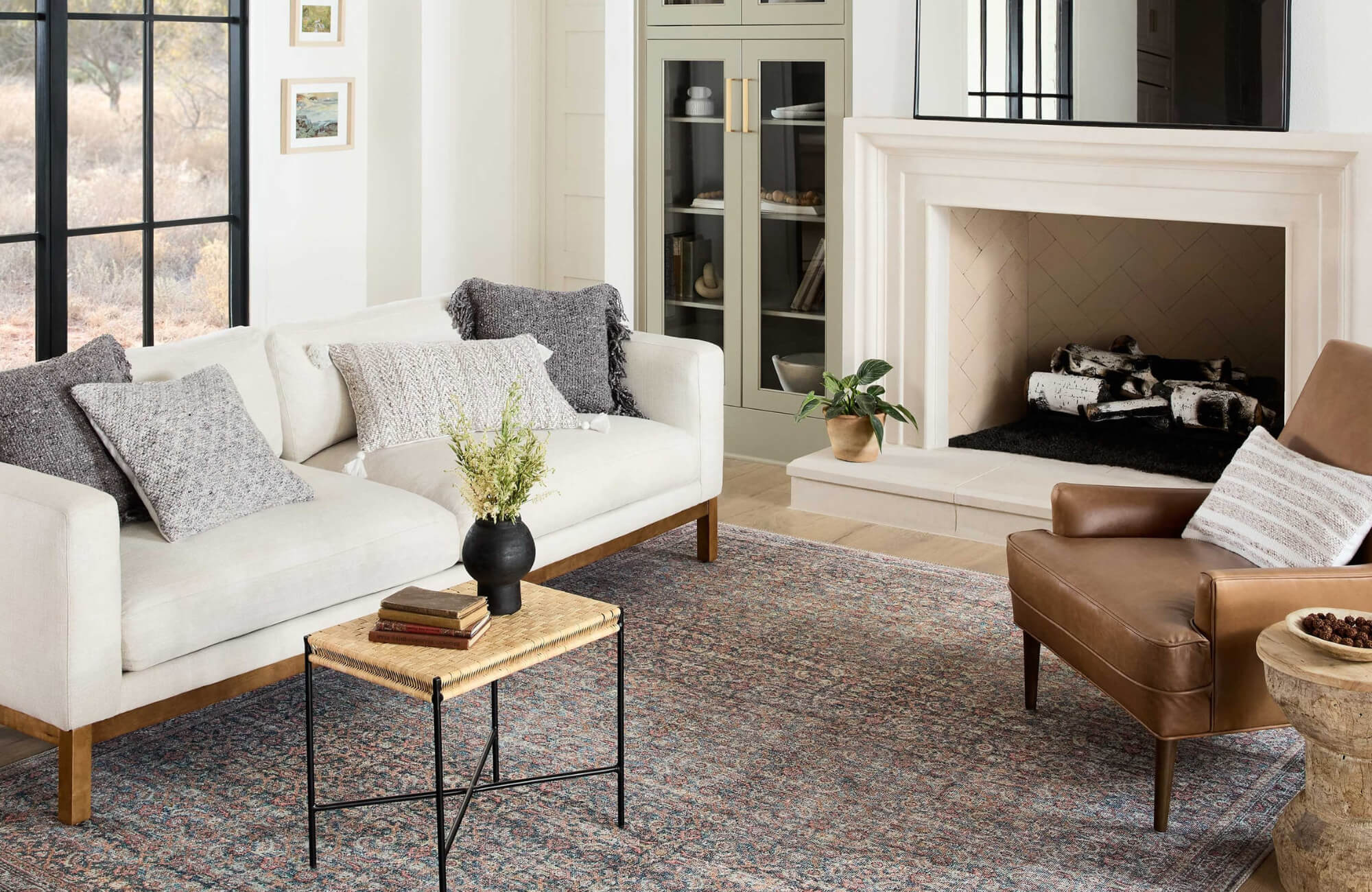 null Bright living room with a cream sofa, leather accent chair, and a muted blue and rust rug, styled near a fireplace and black-framed window.