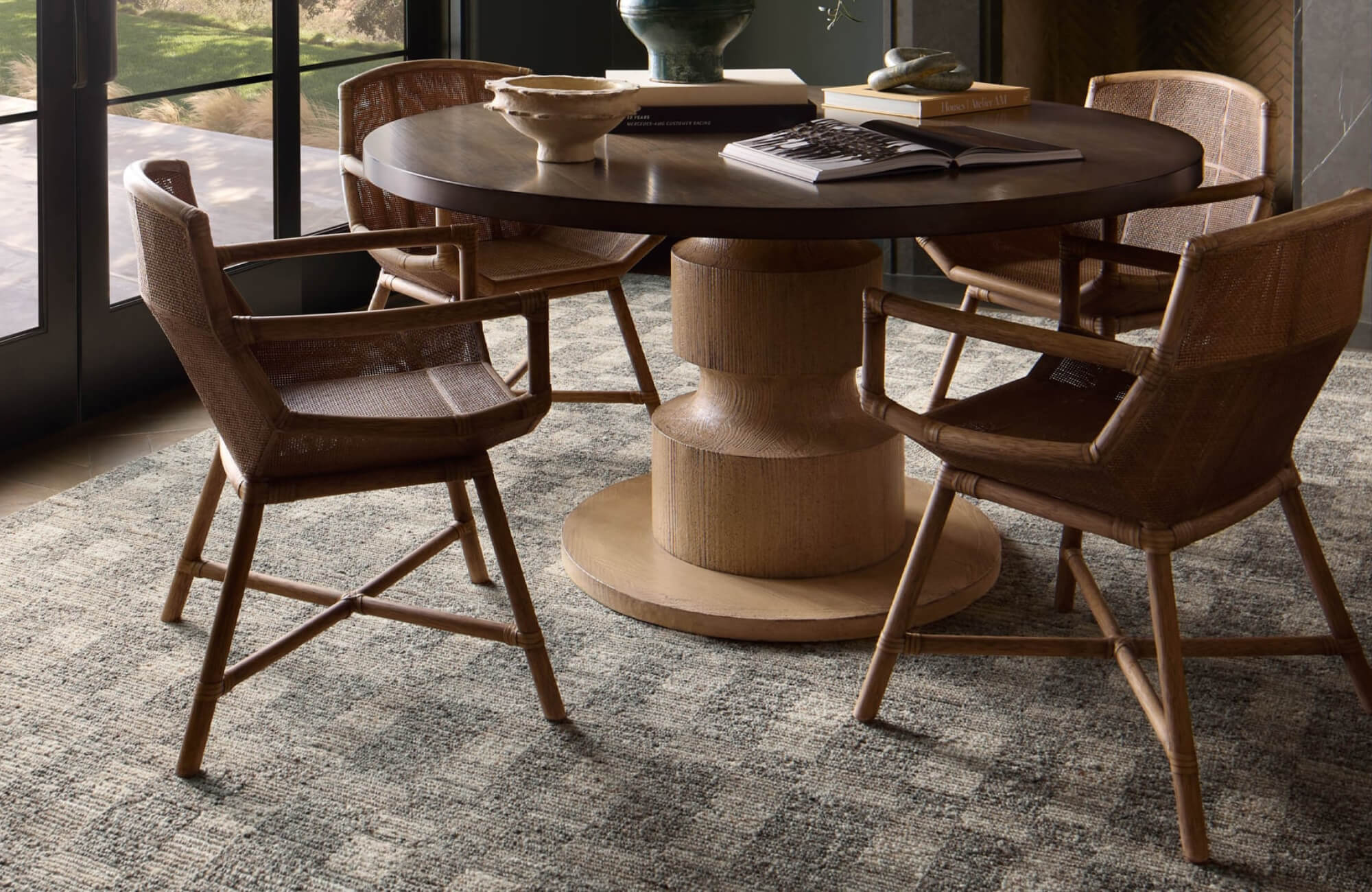 null Dining area with a round wood table, woven chairs, and a textured gray patterned rug near large glass doors overlooking a green yard.