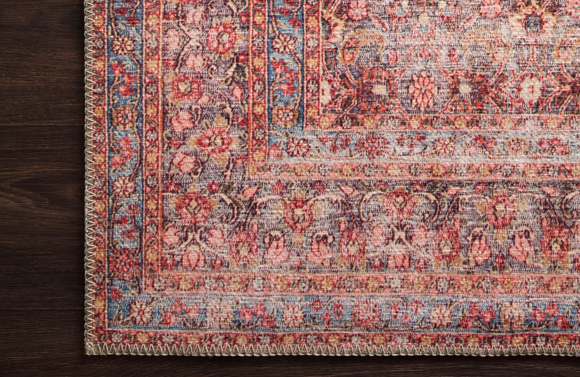 Close-up of a traditional rug corner with intricate floral and vine motifs in soft pink, muted blue, and ivory tones, bordered by a stitched edge.