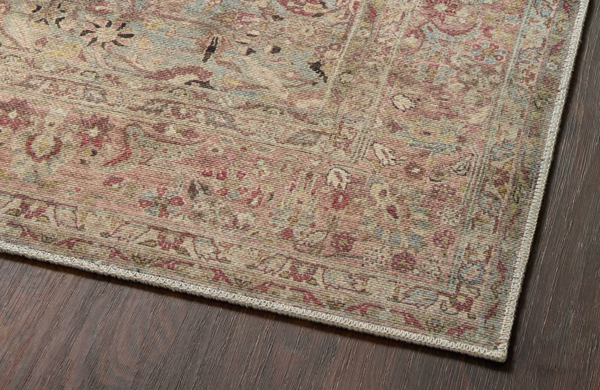 Close-up of a distressed rug corner with faded pink, sage, and beige motifs, featuring a narrow border and stitched edge on dark wood flooring.
