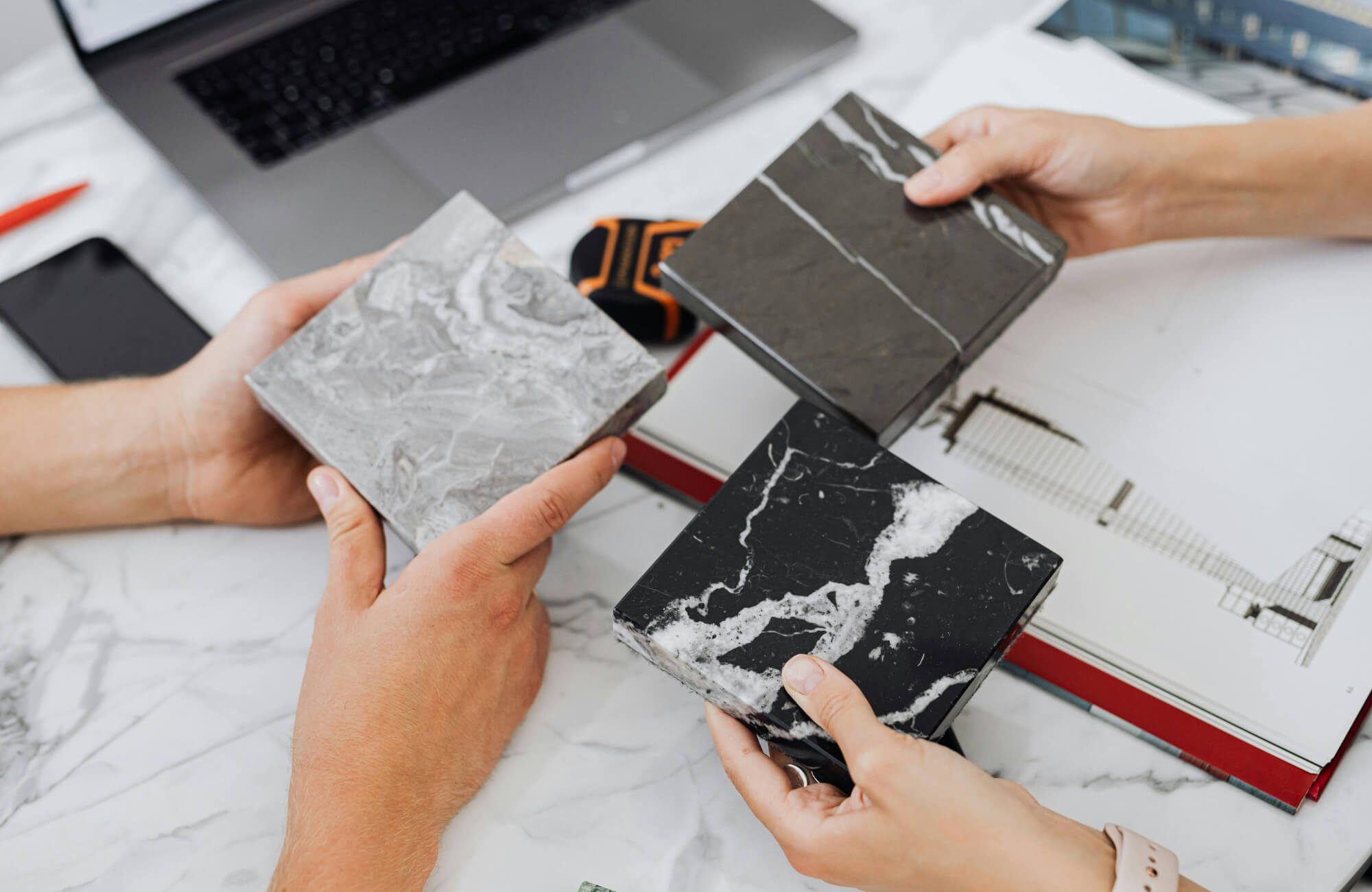 null Two people holding marble-look tile samples in gray, black, and veined finishes over design plans, comparing tones and patterns.