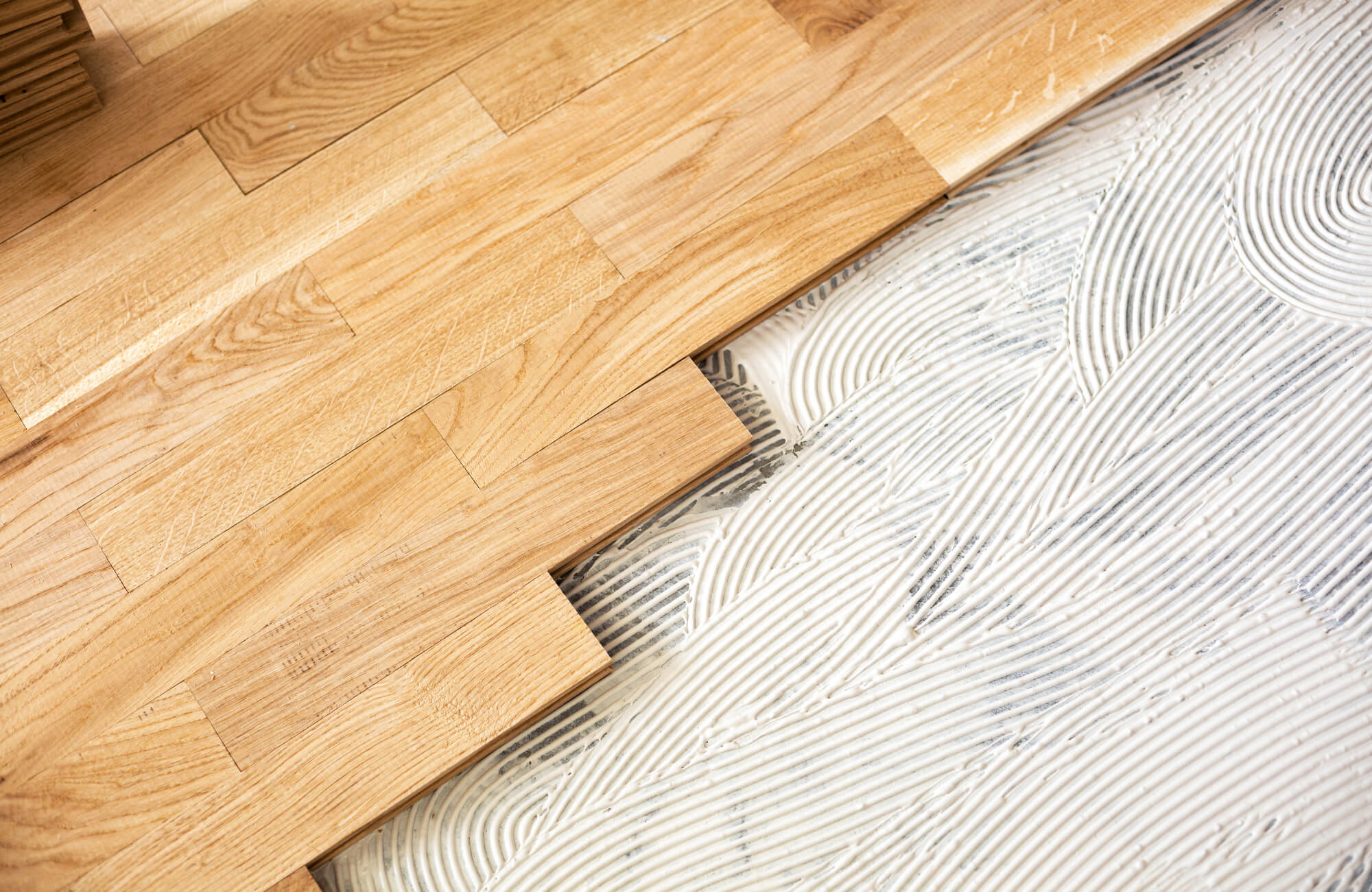 null Hardwood planks installed beside the exposed subfloor with spread adhesive, showing transition preparation between wood flooring and tile base.