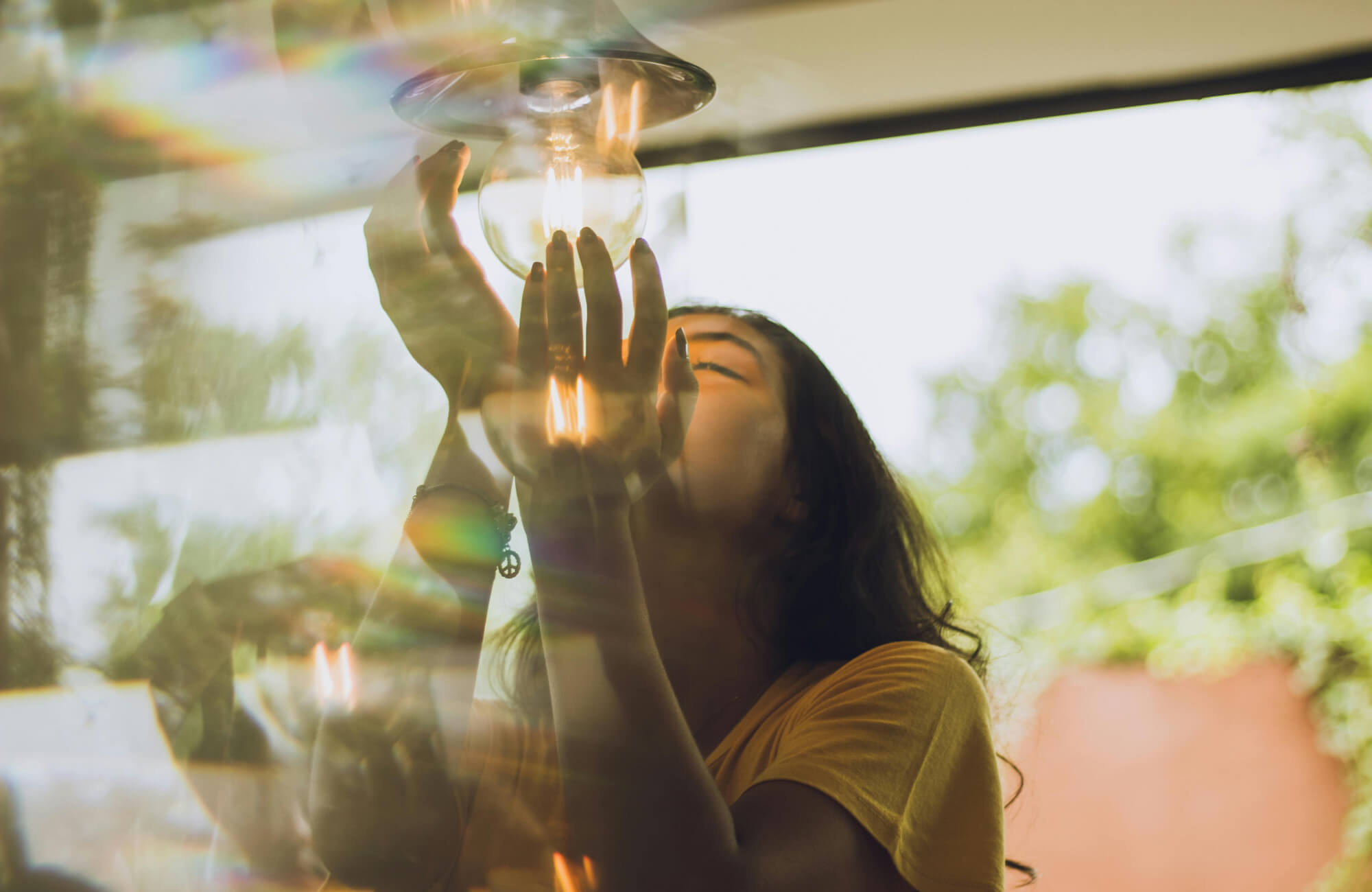 Woman in a yellow shirt adjusting a light bulb indoors, with natural light and rainbow reflections in the background.