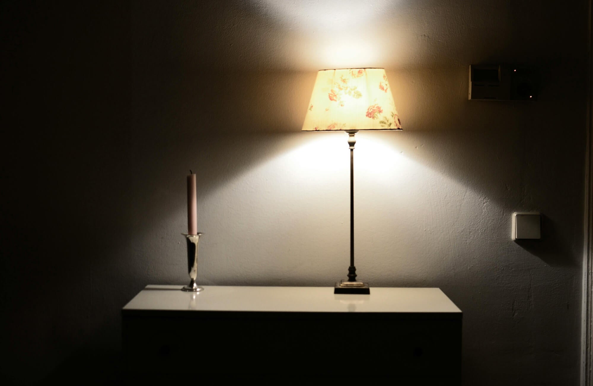 Warmly lit table scene featuring a floral-patterned lamp and a decorative candle in a minimalist, darkened room.
