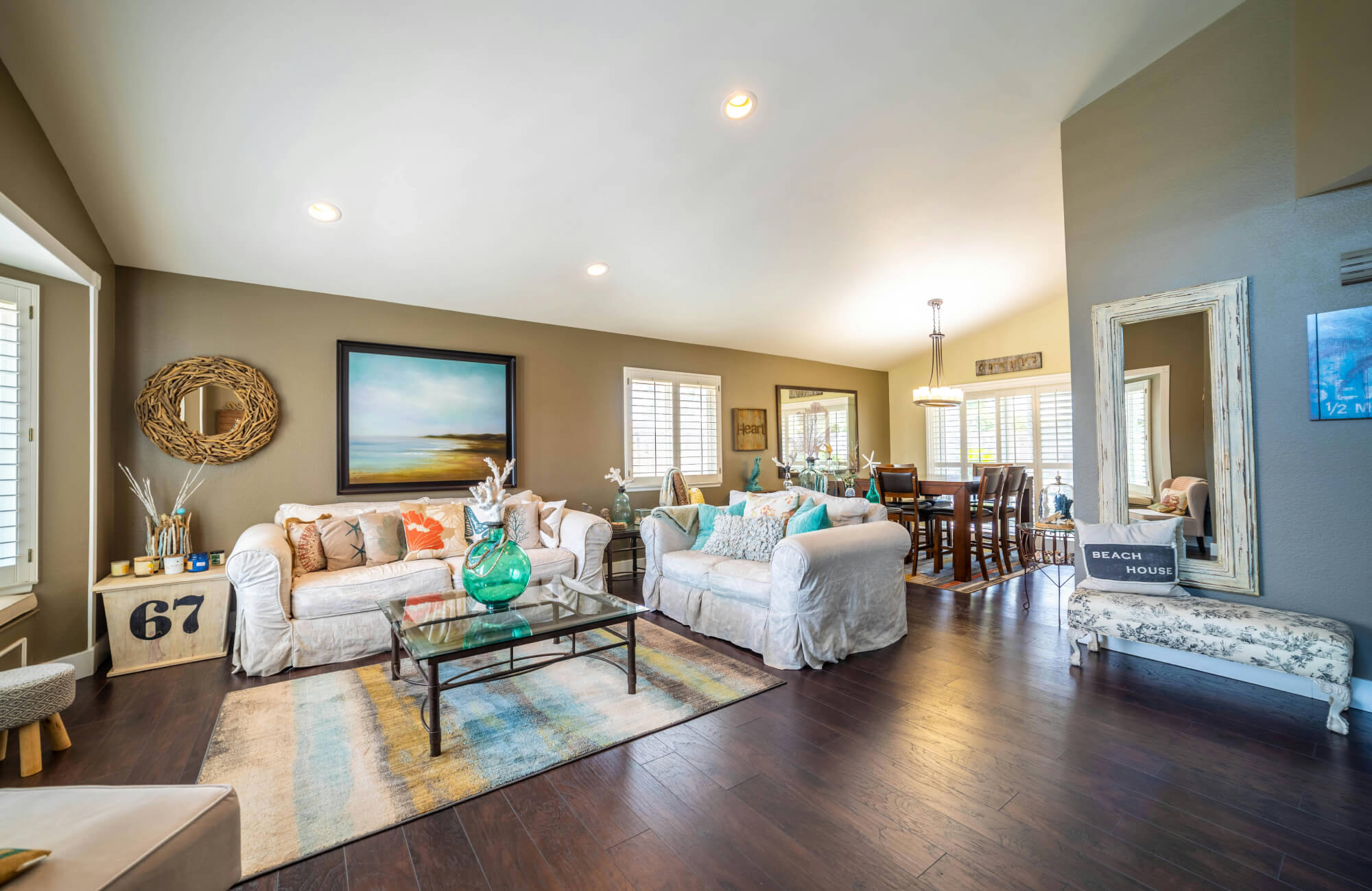 Coastal-inspired living room with dark wood floors, white sofas, beach-themed wall art, and airy open dining area.