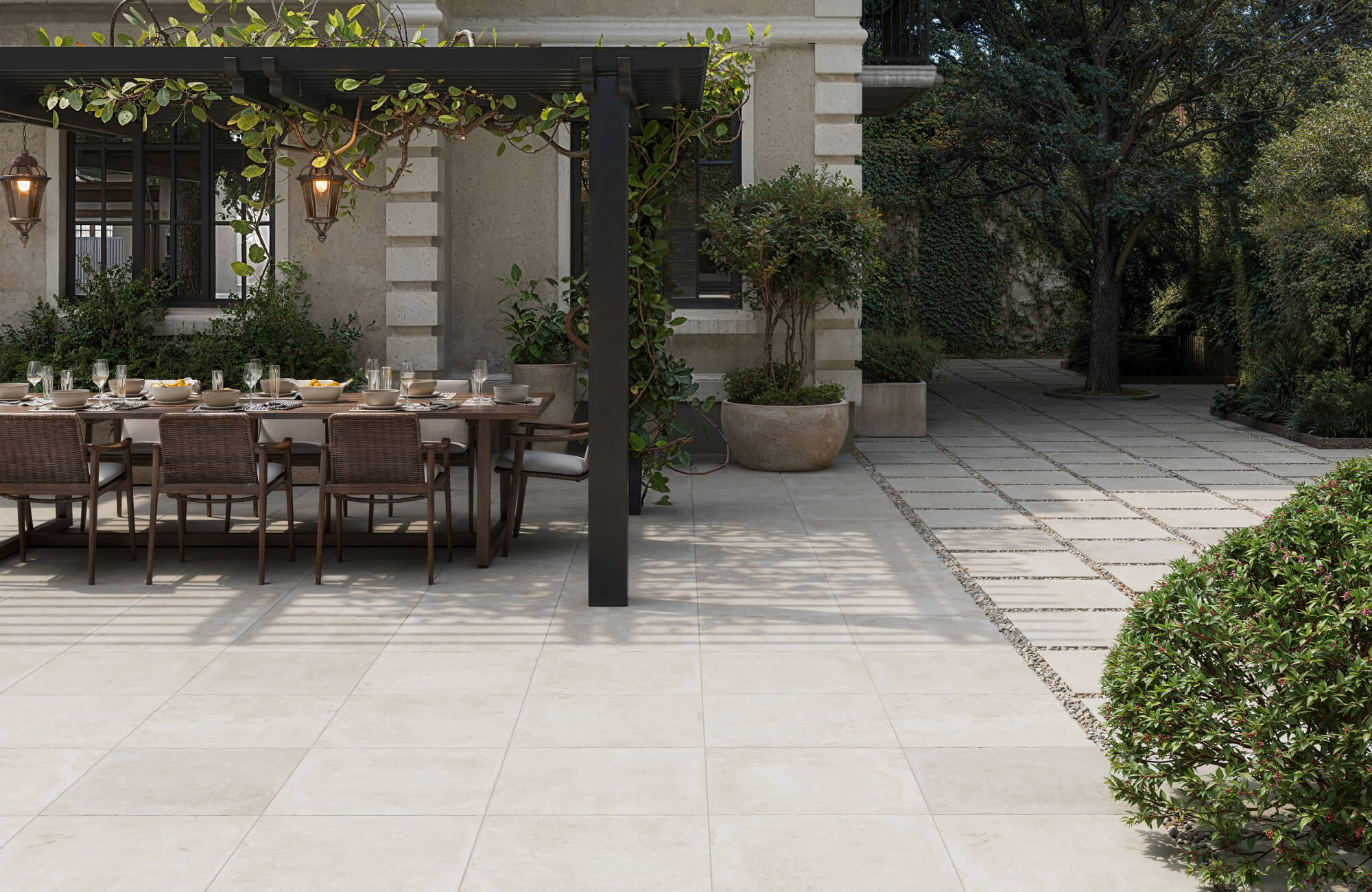 null Elegant outdoor dining area with light paver tile flooring, a pergola draped in greenery, and a stone pathway with pebble accents.