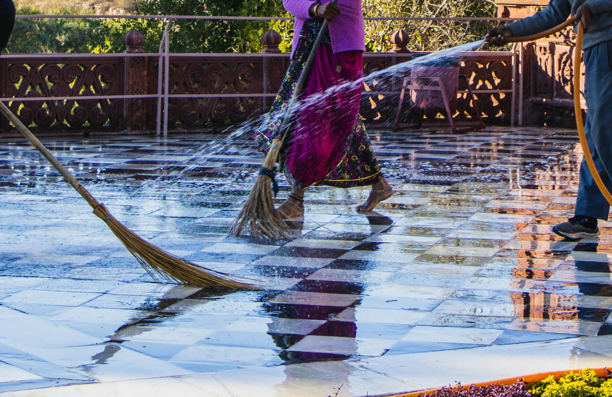 null Workers cleaning a checkered paver tile courtyard with brooms and a hose, reflecting vibrant clothing and ornate red stone railing.