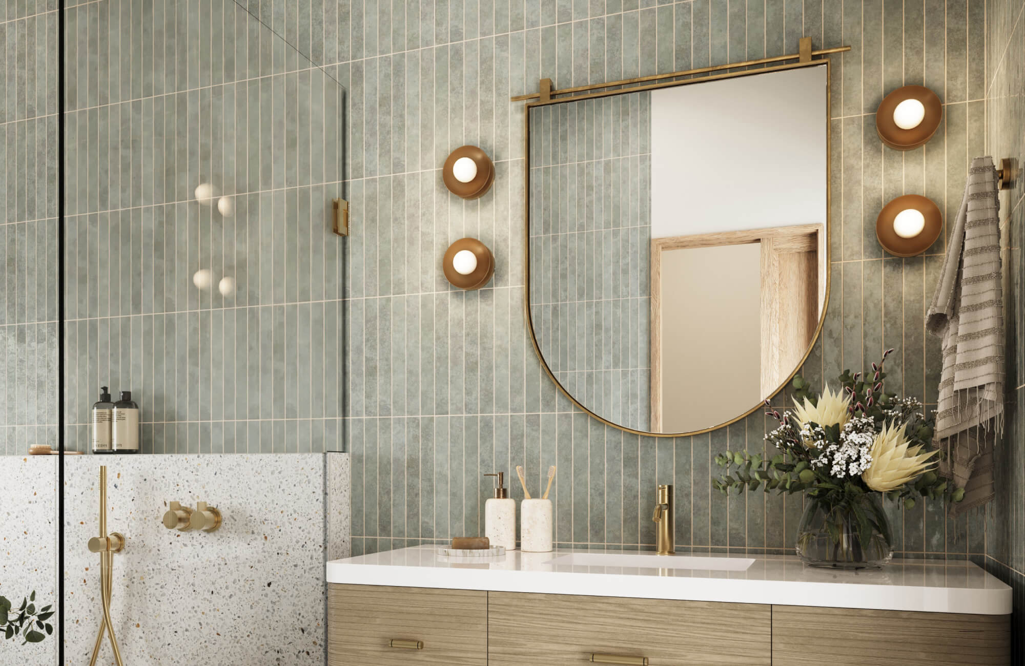 Modern bathroom with a wood vanity, arched brass-framed mirror, vertical sage green tiles, globe sconces, and terrazzo shower surround.