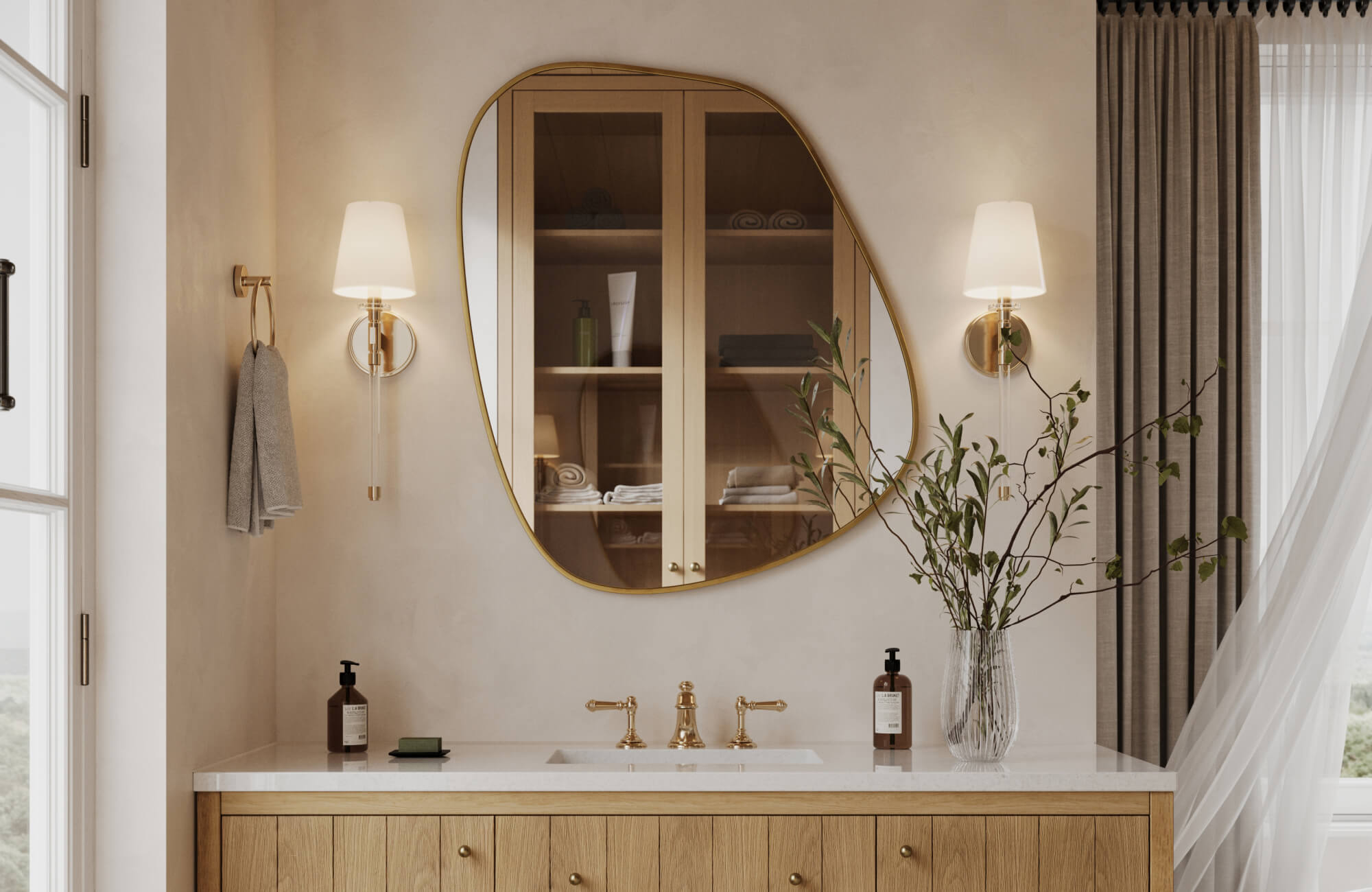 Elegant bathroom with a light wood vanity, sculptural brass-framed mirror, and tapered wall sconces with white shades.