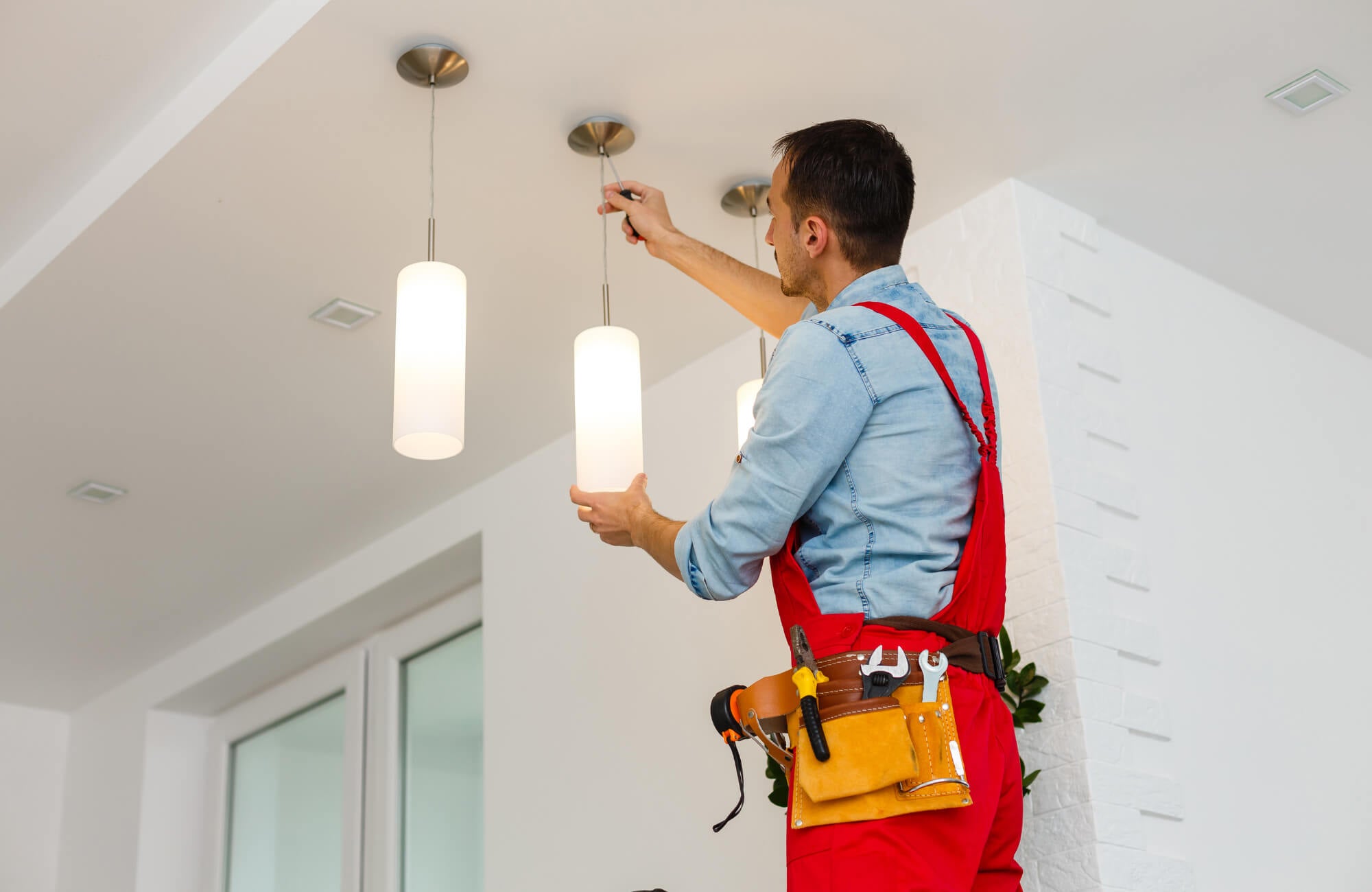 null Electrician in red overalls installing cylindrical pendant lights on a white ceiling inside a bright modern kitchen.
