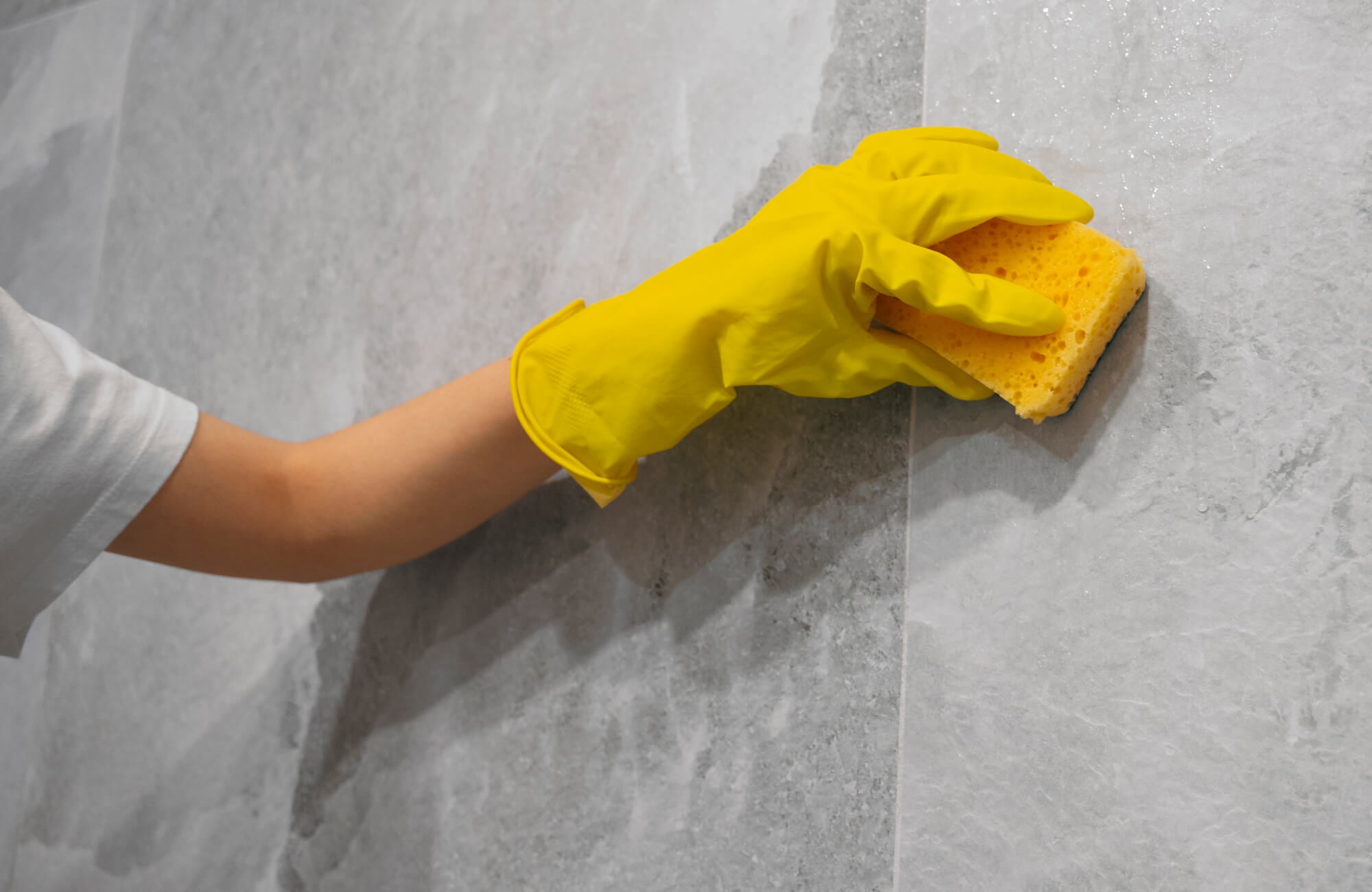 null Hand in yellow glove cleaning a gray cement look porcelain tile wall with a sponge, highlighting easy tile maintenance.