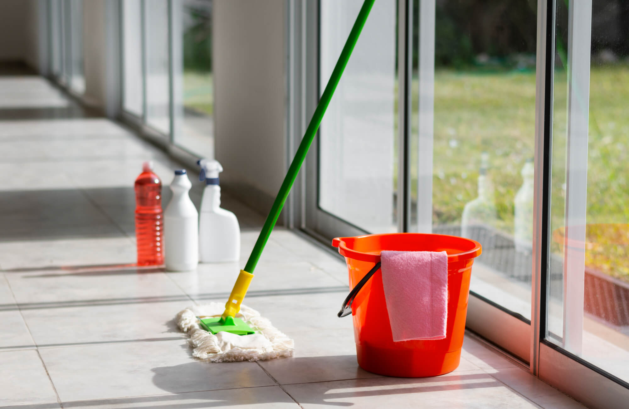 Porcelain tile cleaning essentials including mop, bucket, and spray bottles arranged on a sunlit tiled floor.