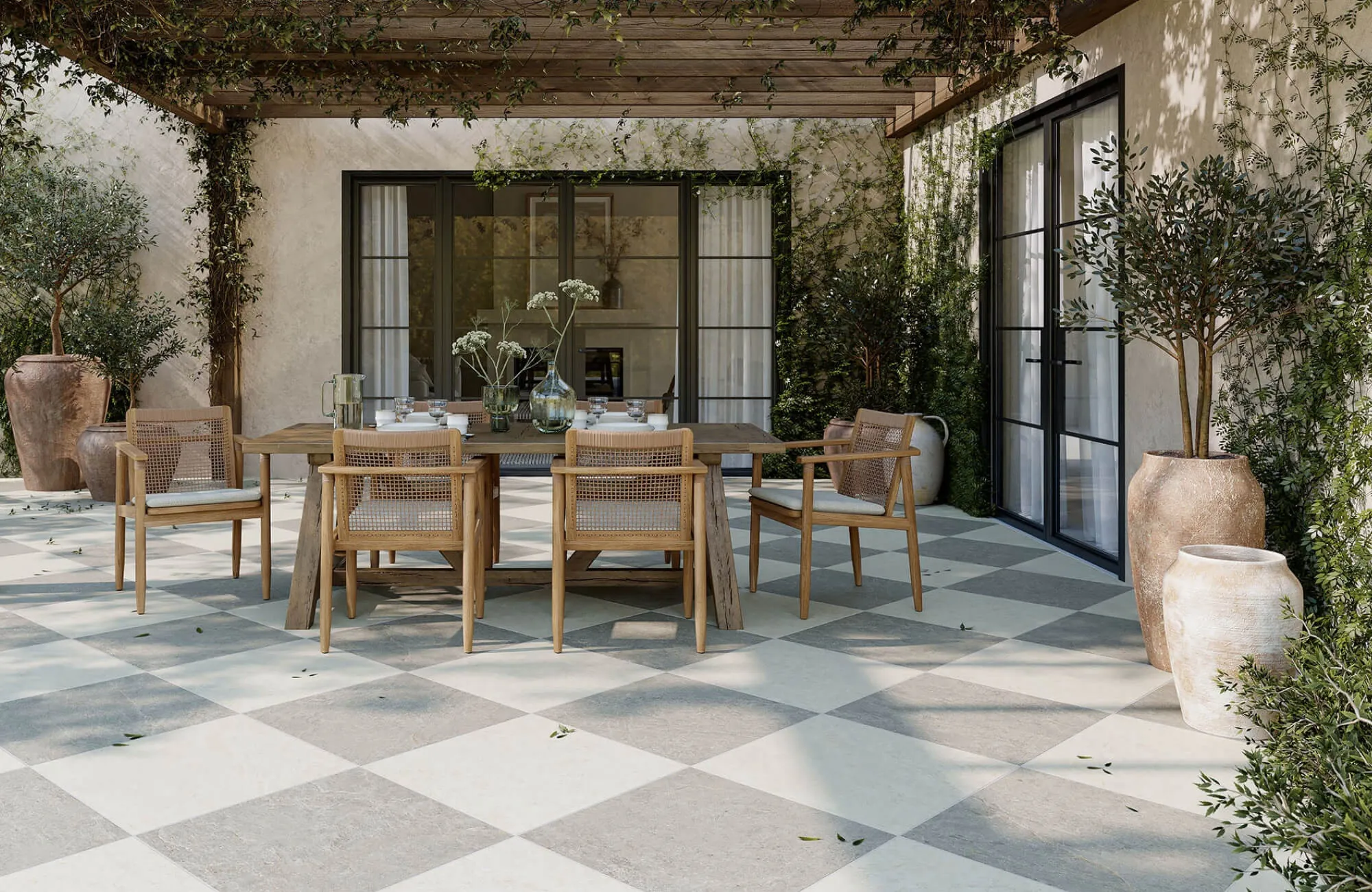 Outdoor patio dining area with a rectangular wooden table, woven armchairs, large planters, glass doors, and a shaded pergola covered in greenery.