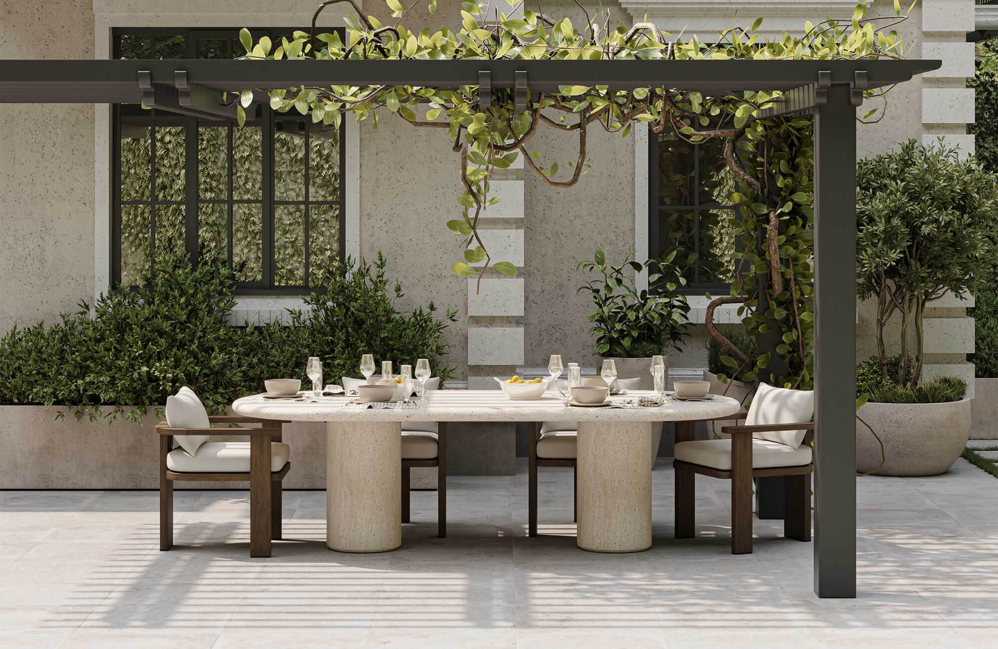 Outdoor dining area beneath a pergola with an oval stone table, cushioned wooden chairs, greenery, potted plants, and a softly styled tablescape.