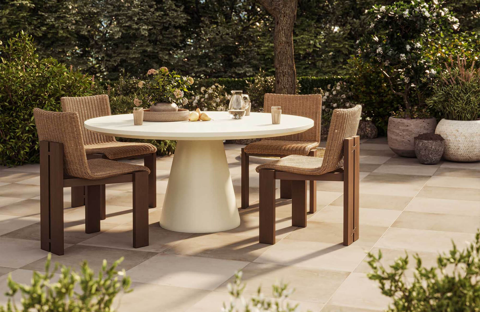 Garden patio with a round pedestal dining table, woven chairs, tabletop drinks and decor, surrounding planters, and trees providing natural shade.